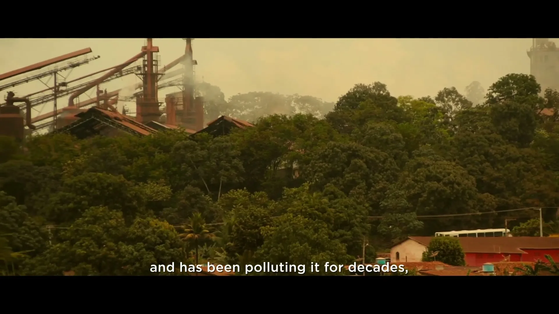 A wide shot shows a small town nestled among lush green trees, with a large, industrial steel plant looming in the background, emitting smoke into a hazy sky.