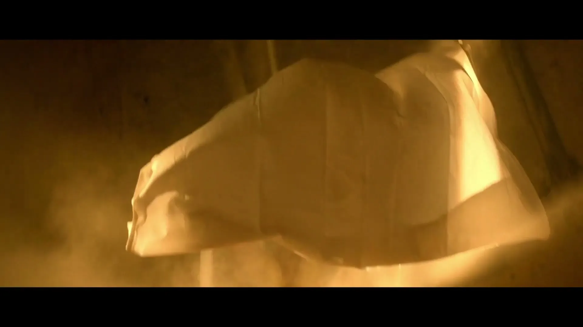 A white sheet or cloth billows and falls in a dusty, dimly lit room, creating a sense of mystery and movement.