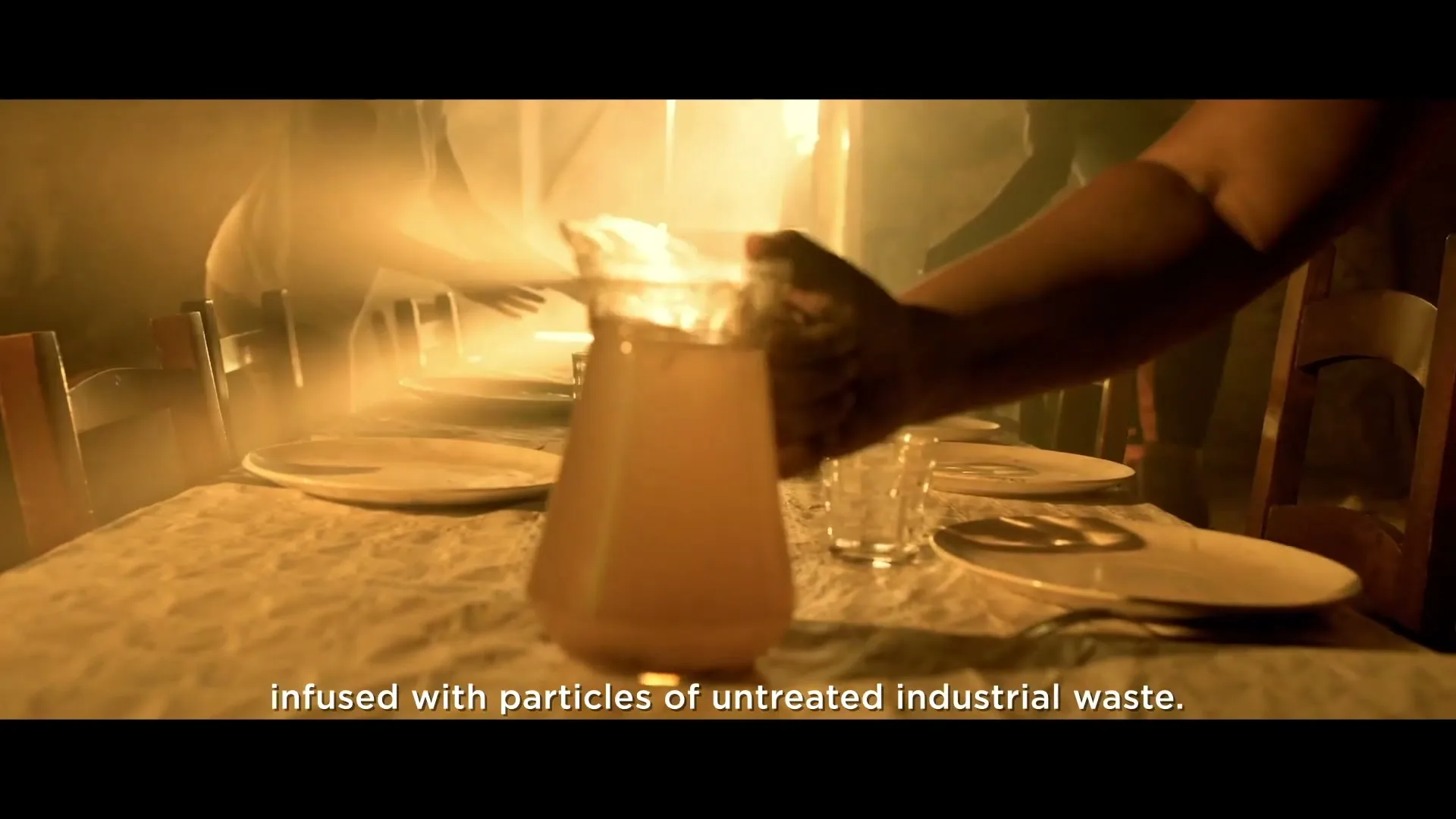 A person places a pitcher of murky water on a table set with plates and glasses, in a dimly lit, smoky room.
