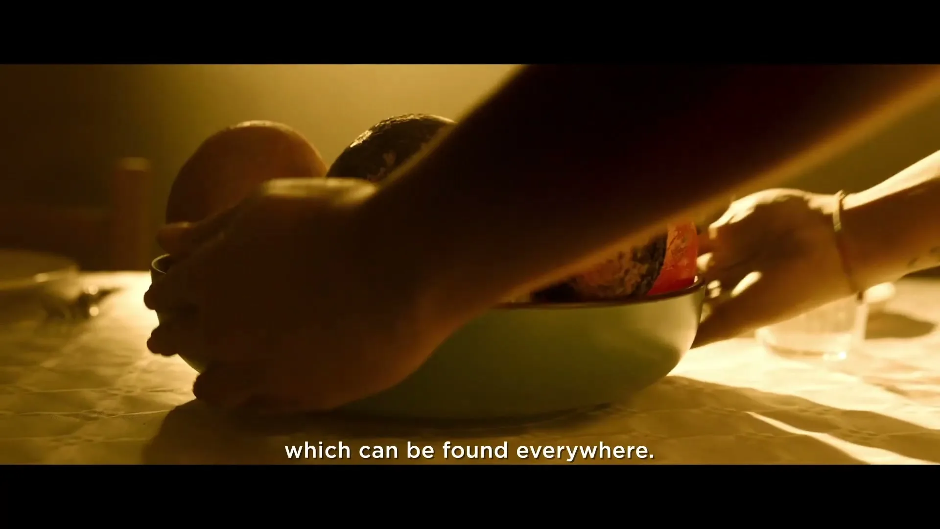 A person's hands place a bowl of fruit, some of which appears rotten or covered in dust, onto a table with a white tablecloth in a dimly lit room.
