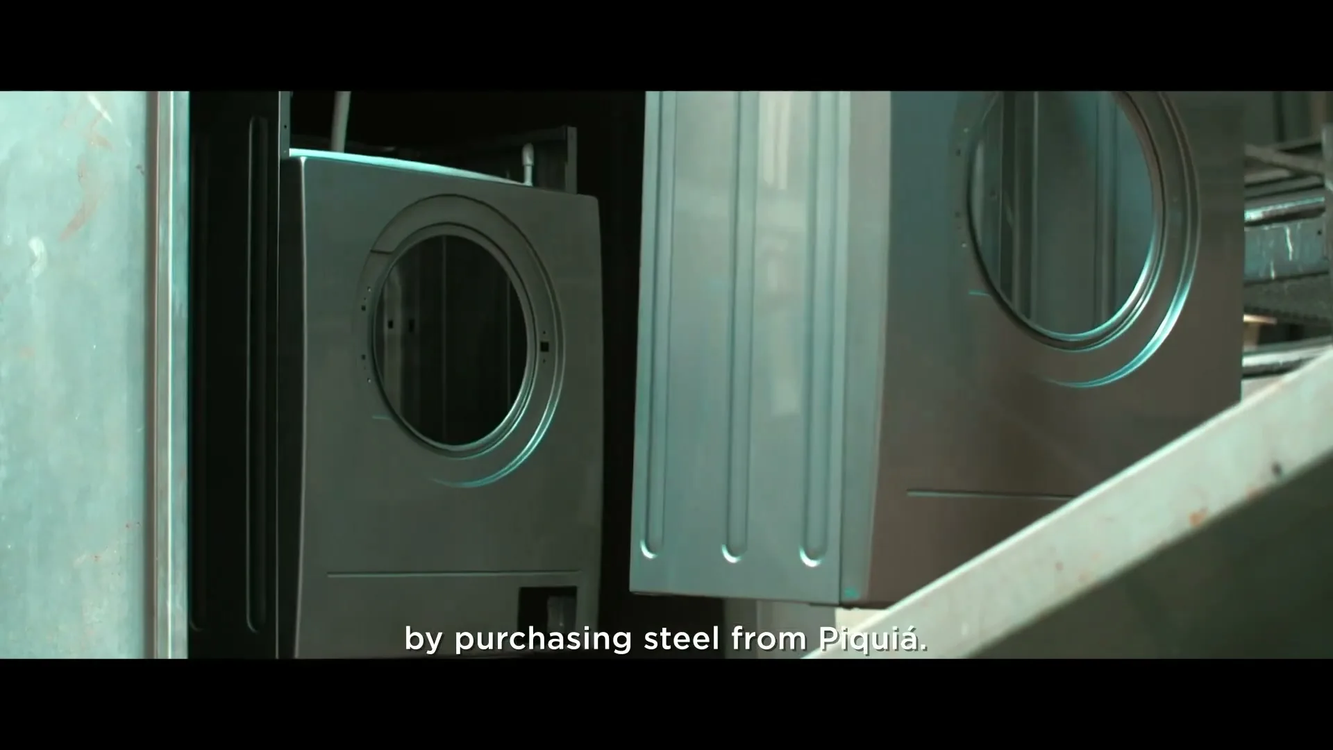 Two metal washing machine shells move along a conveyor belt in a dimly lit factory, with text overlayed on the screen.