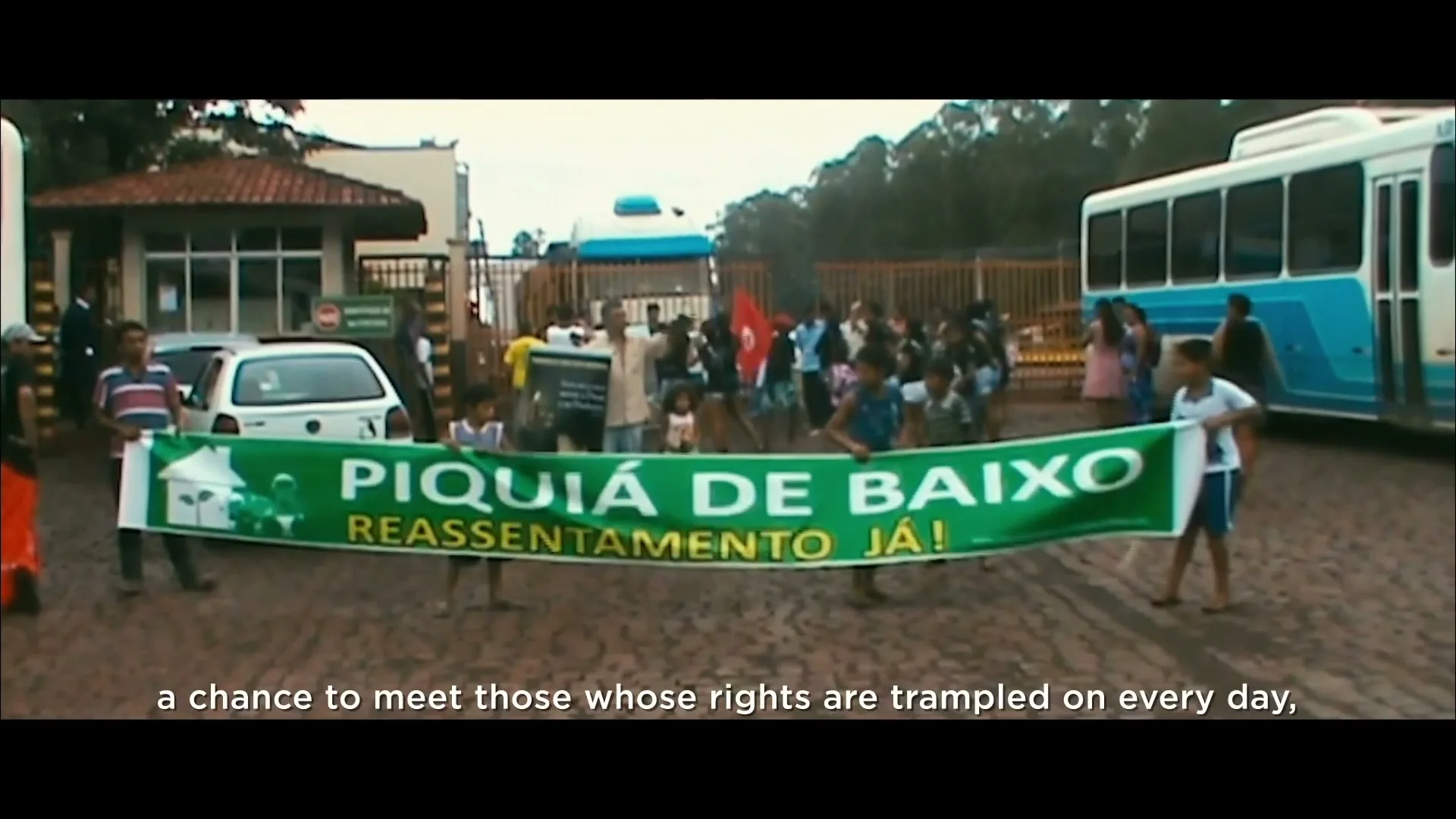 A group of people, including children, hold a large green banner that reads "PIQUIÁ DE BAIXO REASSENTAMENTO JÁ!" at an outdoor protest or gathering.