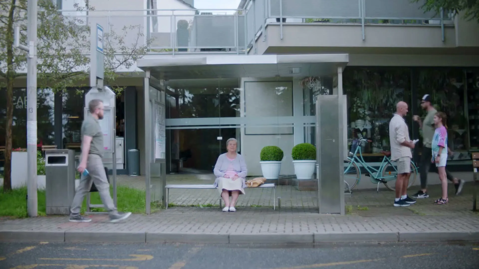An elderly woman sits alone at a bus stop while people walk by and a car drives past, on a cloudy day in a suburban area.