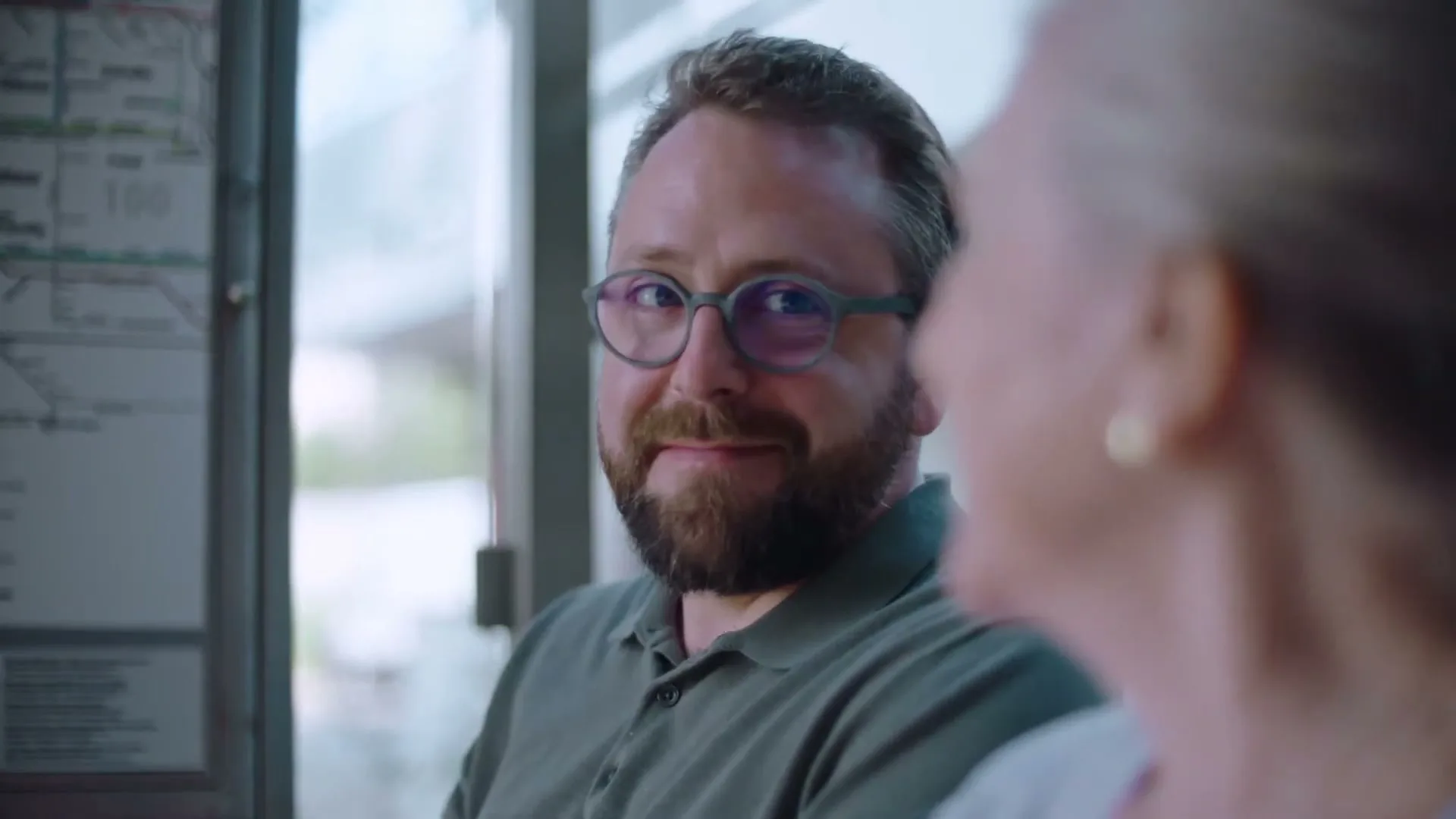 A man with a beard and glasses looks at a woman next to him and smiles while sitting at a bus stop.