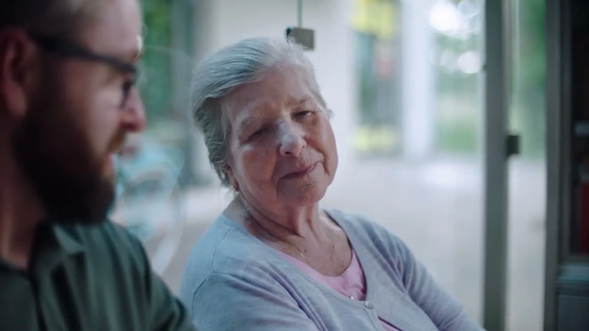 An older woman with grey hair and a younger man with a beard sit together at a bus stop, looking at each other and smiling.