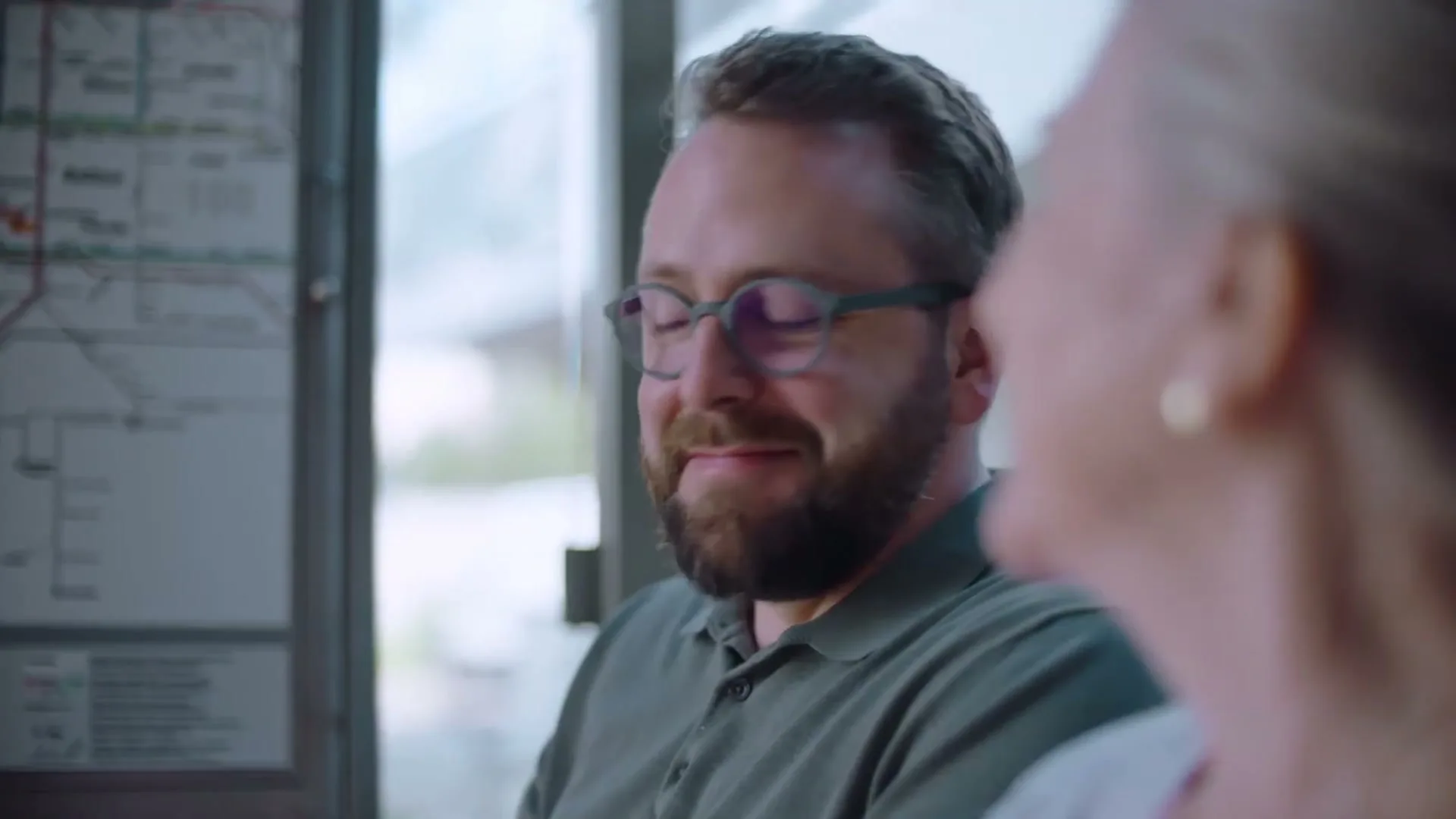 A bearded man in glasses smiles at an older woman, sitting together at a bus stop during the day.