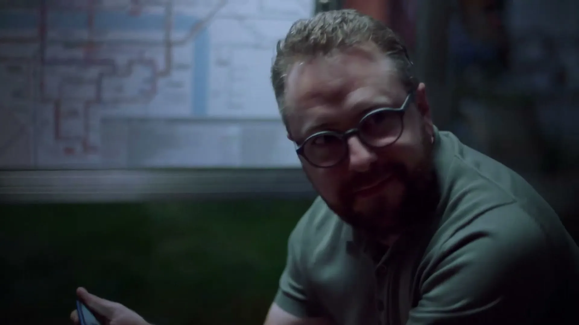 A man with a beard and glasses looks up from his phone and smiles, then reacts in surprise at a bus stop at night.