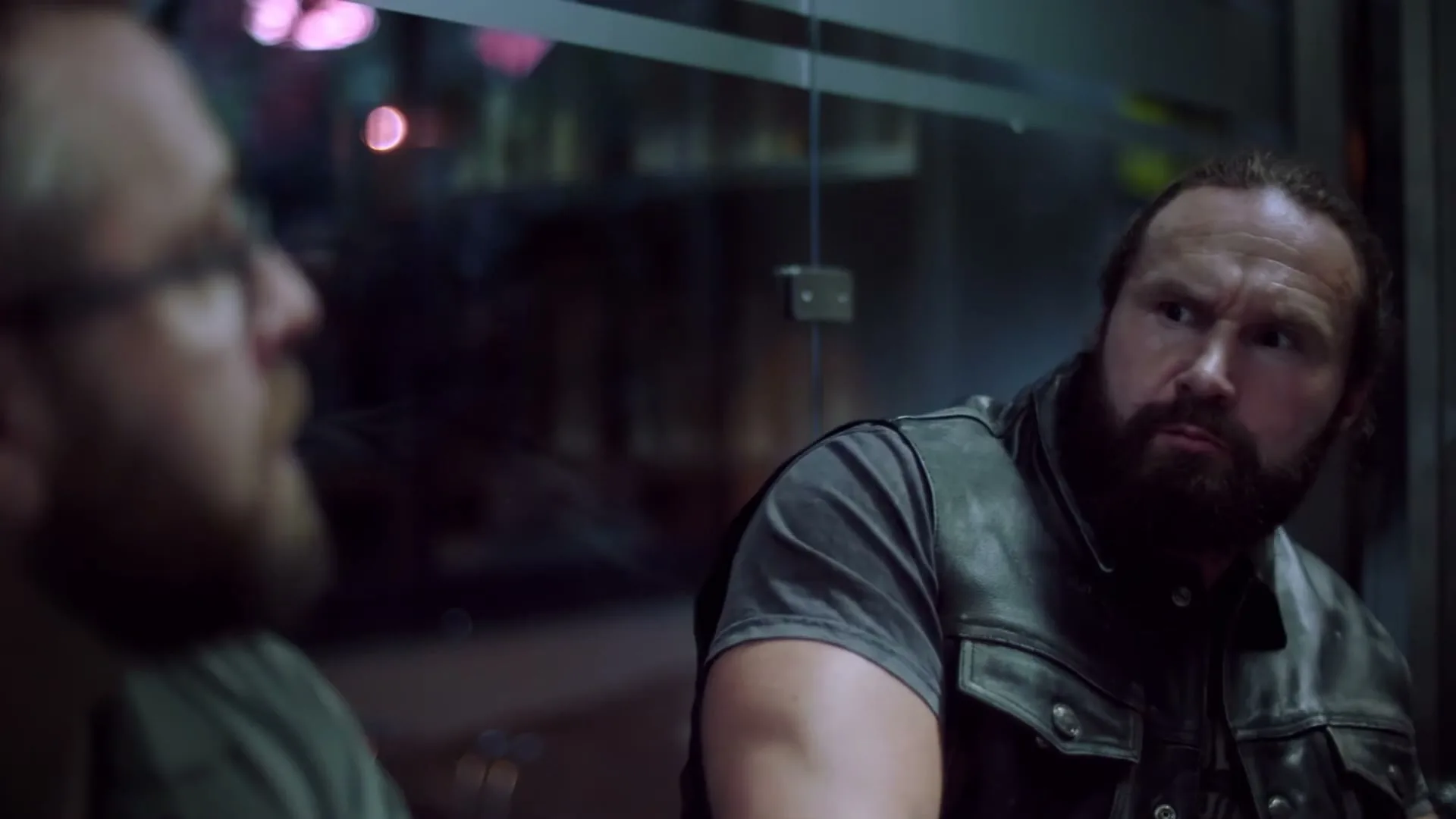 A burly man with a beard and a leather vest looks intently at another man in glasses, who is out of focus in the foreground, inside a building at night.