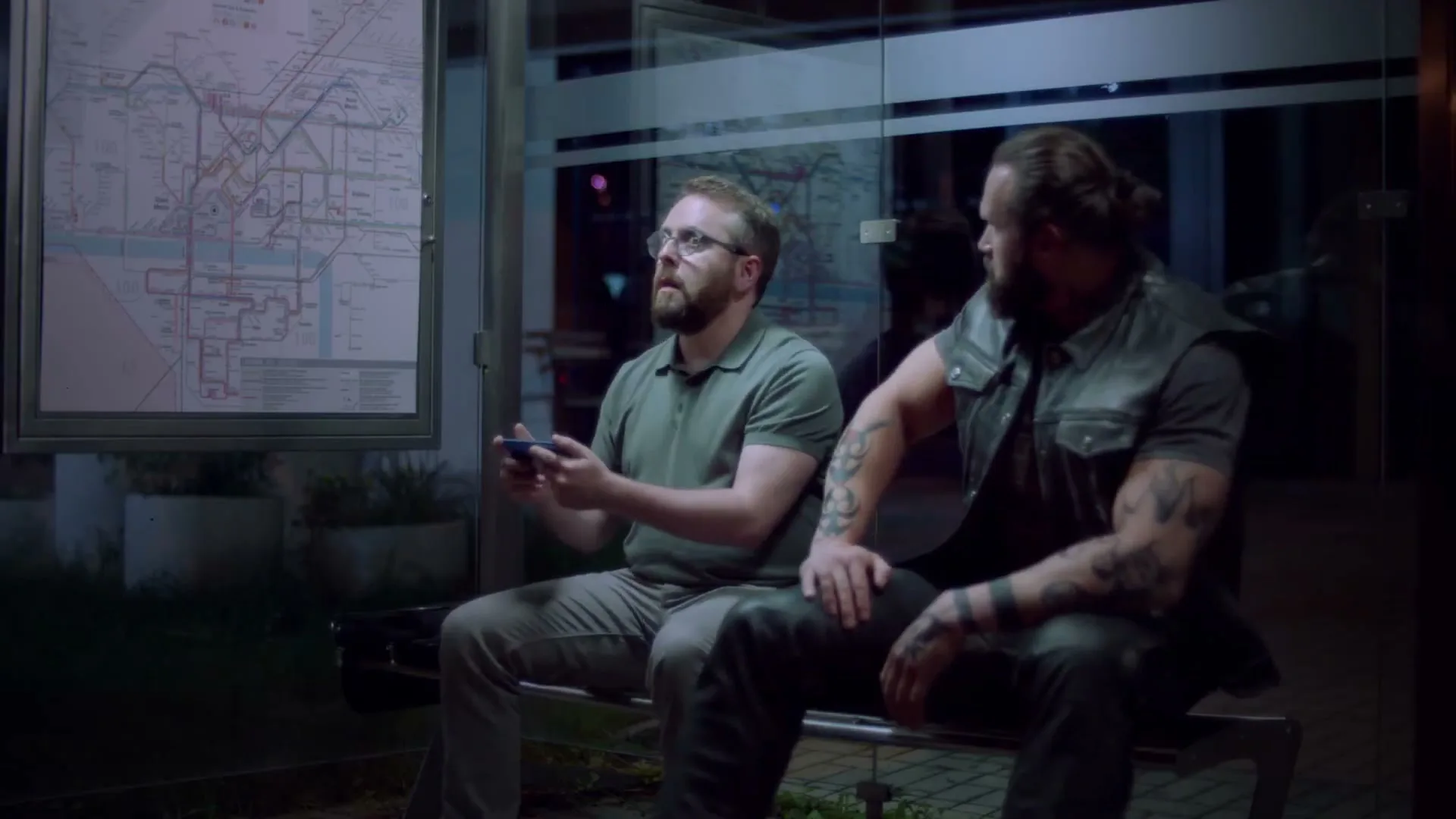 A man with glasses plays a video game on his phone while sitting at a bus stop at night, next to a large, tattooed man.