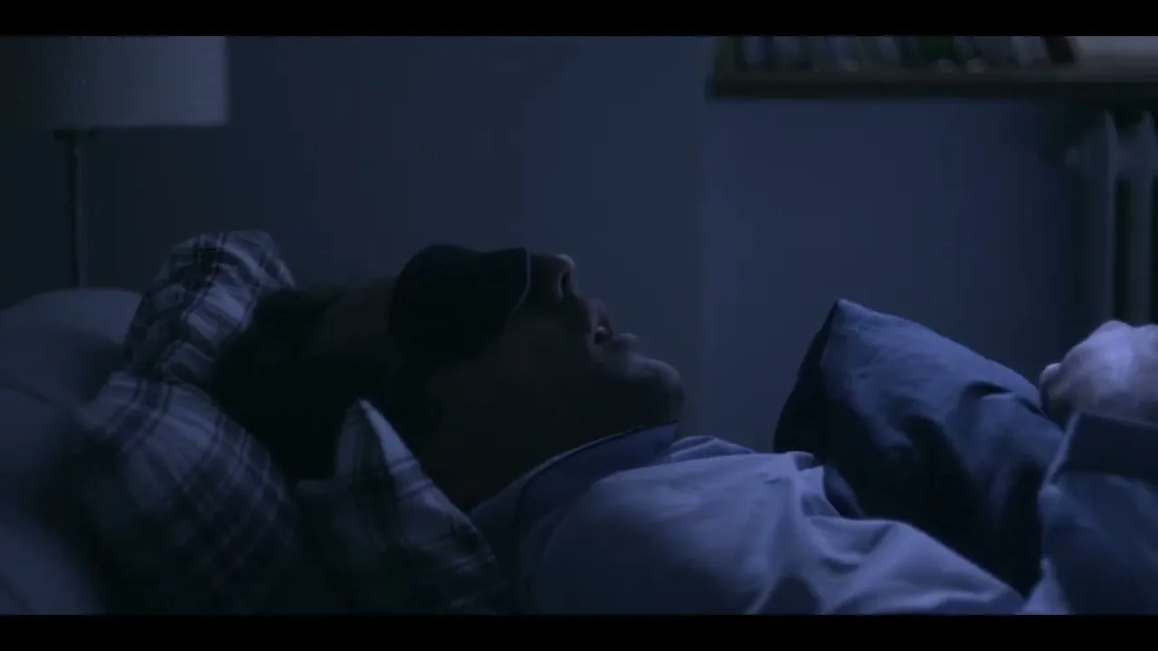 A man in his 30s, wearing a sleep mask, wakes up suddenly in bed at night, looking startled and confused in his bedroom.