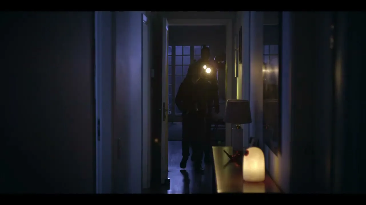 Two burglars, dressed in dark clothing, walk down a dark hallway, illuminating their path with flashlights, in a house at night.