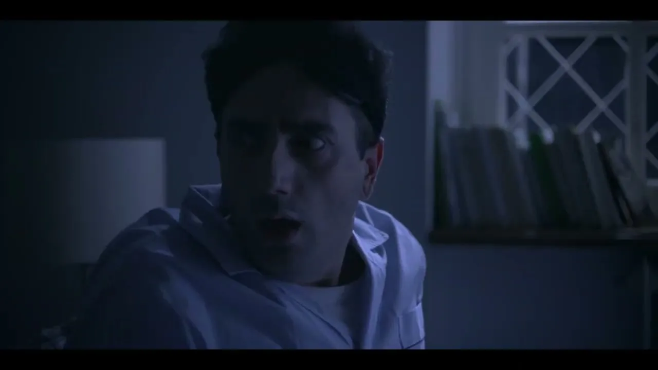 A man with messy hair and a sleep mask on his forehead wakes up in bed, looking shocked and scared in a dark room.