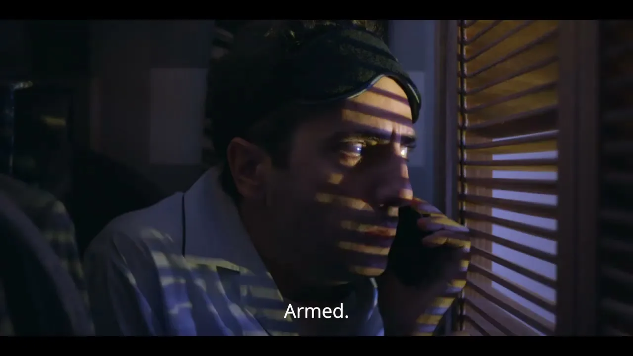 A man in his 30s, wearing pajamas and a sleep mask, looks out a window through blinds while holding a phone to his ear at night.