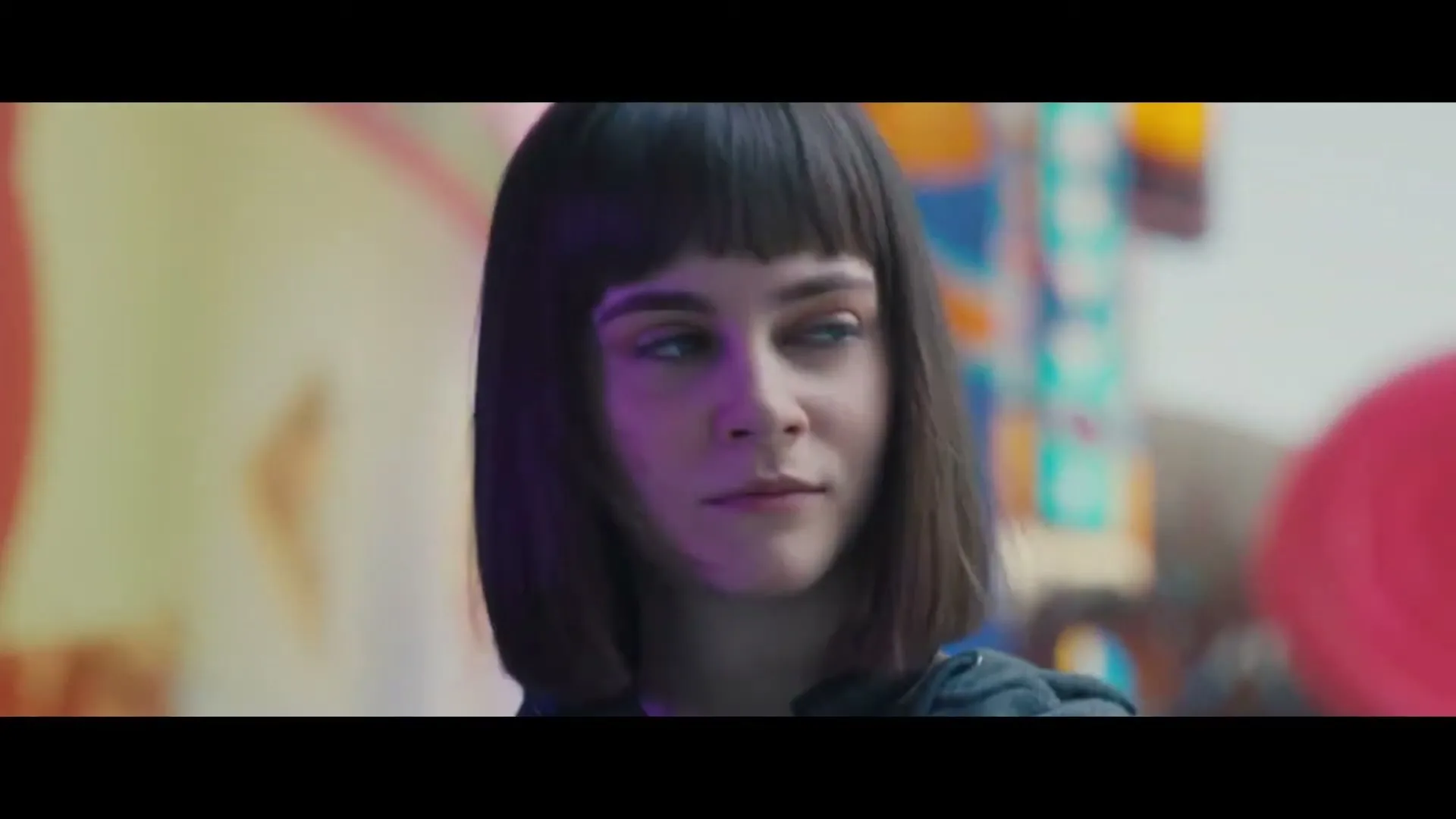 A young woman with a bob haircut and bangs looks up and to the right, with colorful blurred shapes in the background.