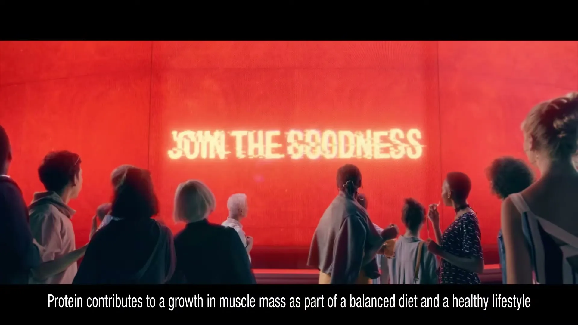 A diverse group of people stand in front of a large red screen that displays the text "JOIN THE GOODNESS" in a stylized font.