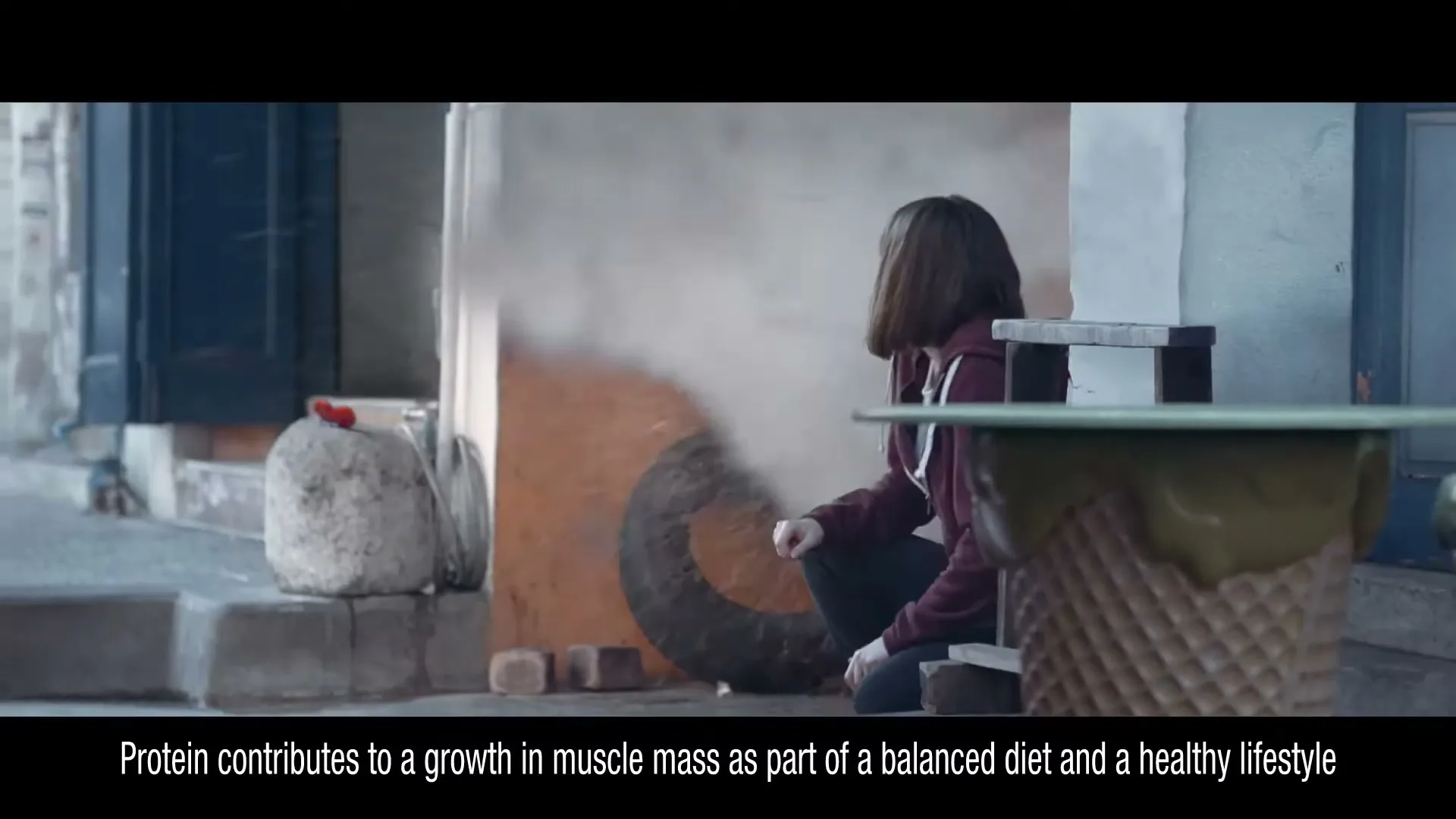 A young woman in a hoodie crouches by a wall, then flinches as an explosion of dust and debris erupts behind her in an alleyway.