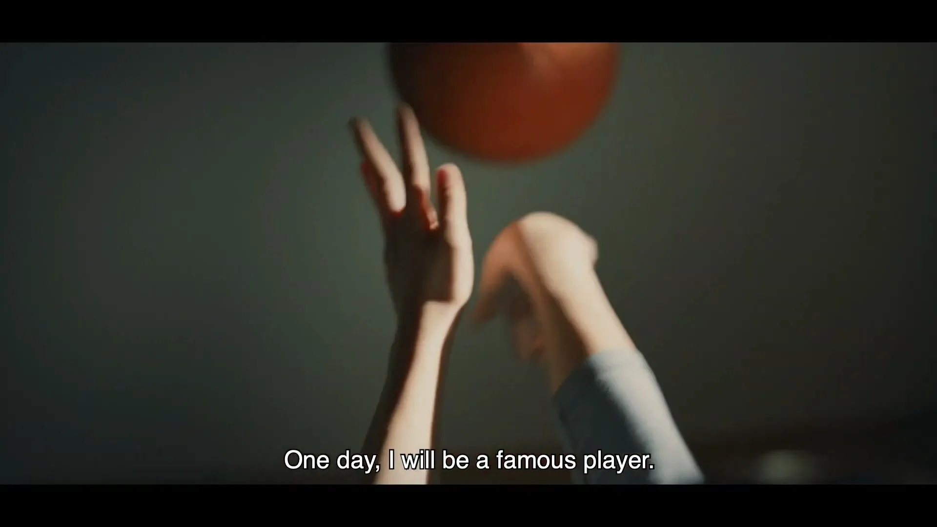 A child's hands are seen bouncing an orange basketball in a dimly lit room, with sunlight streaming in from the left.