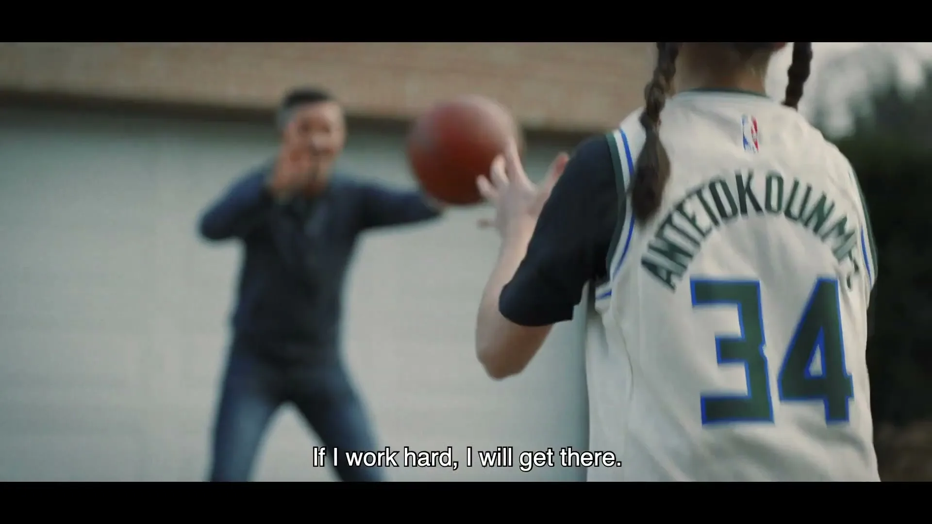 A young girl in a basketball jersey plays with a man in a driveway, catching a basketball.