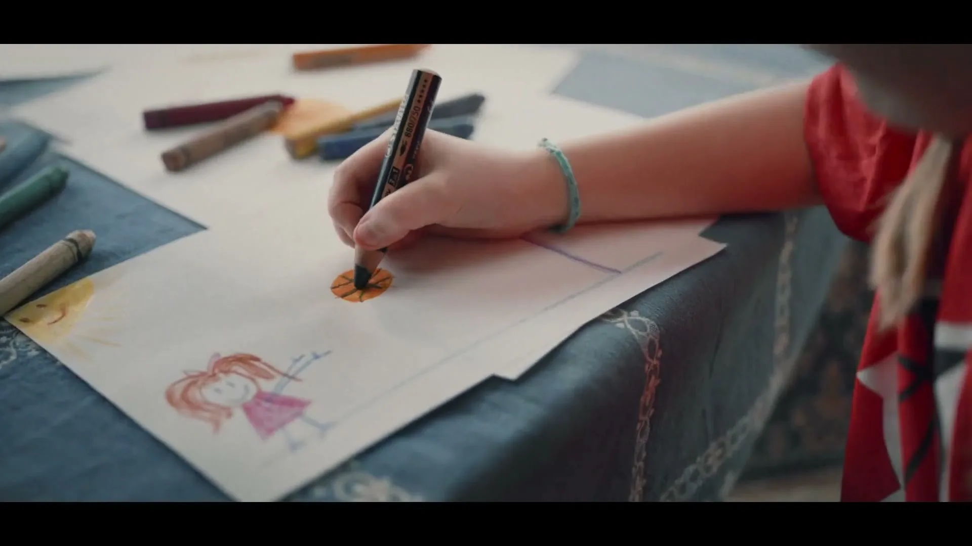 A child's hand draws a basketball on a piece of paper, next to a drawing of a girl, on a blue tablecloth indoors.