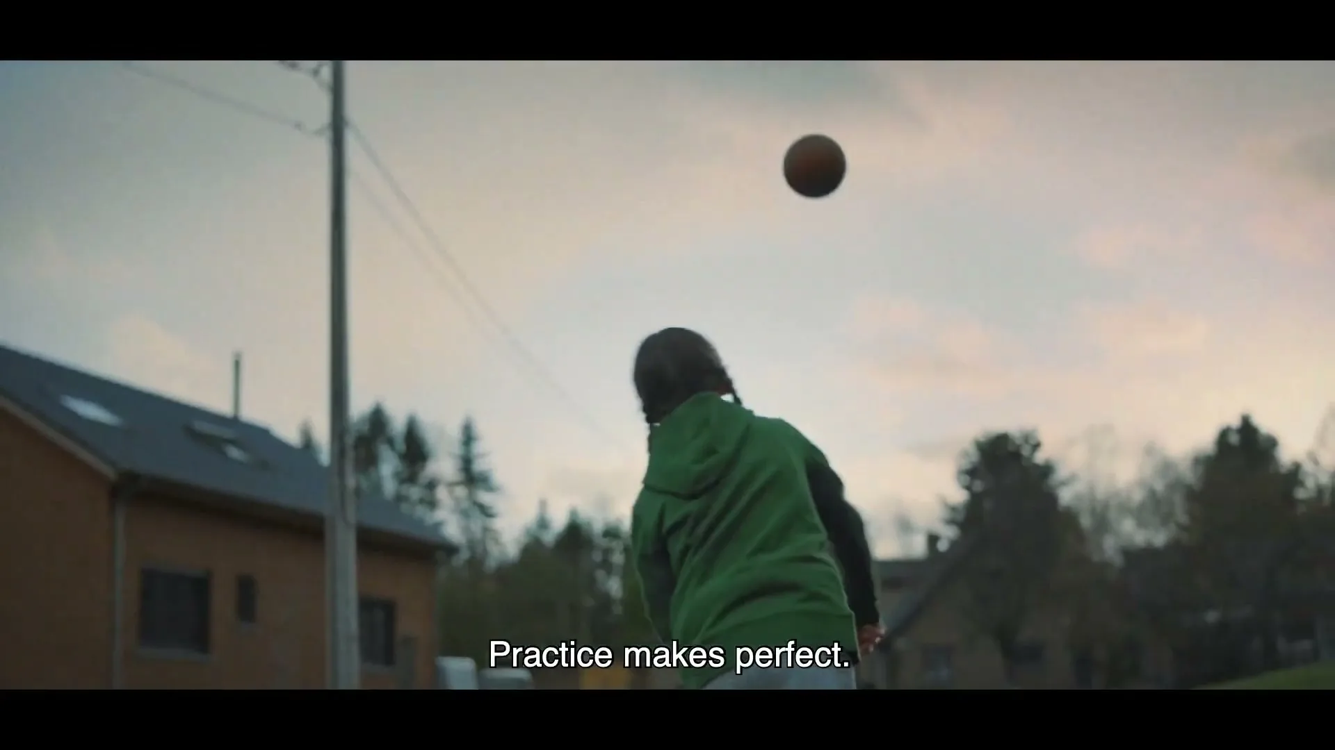 A young girl in a green hoodie throws a basketball into the air, practicing alone on a quiet suburban street at dusk.
