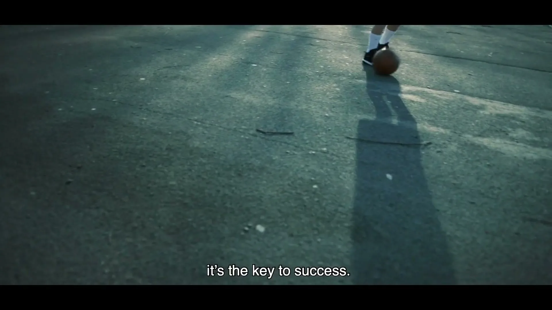 A person's feet dribble a basketball on a cracked asphalt court, with sunlight casting long shadows.