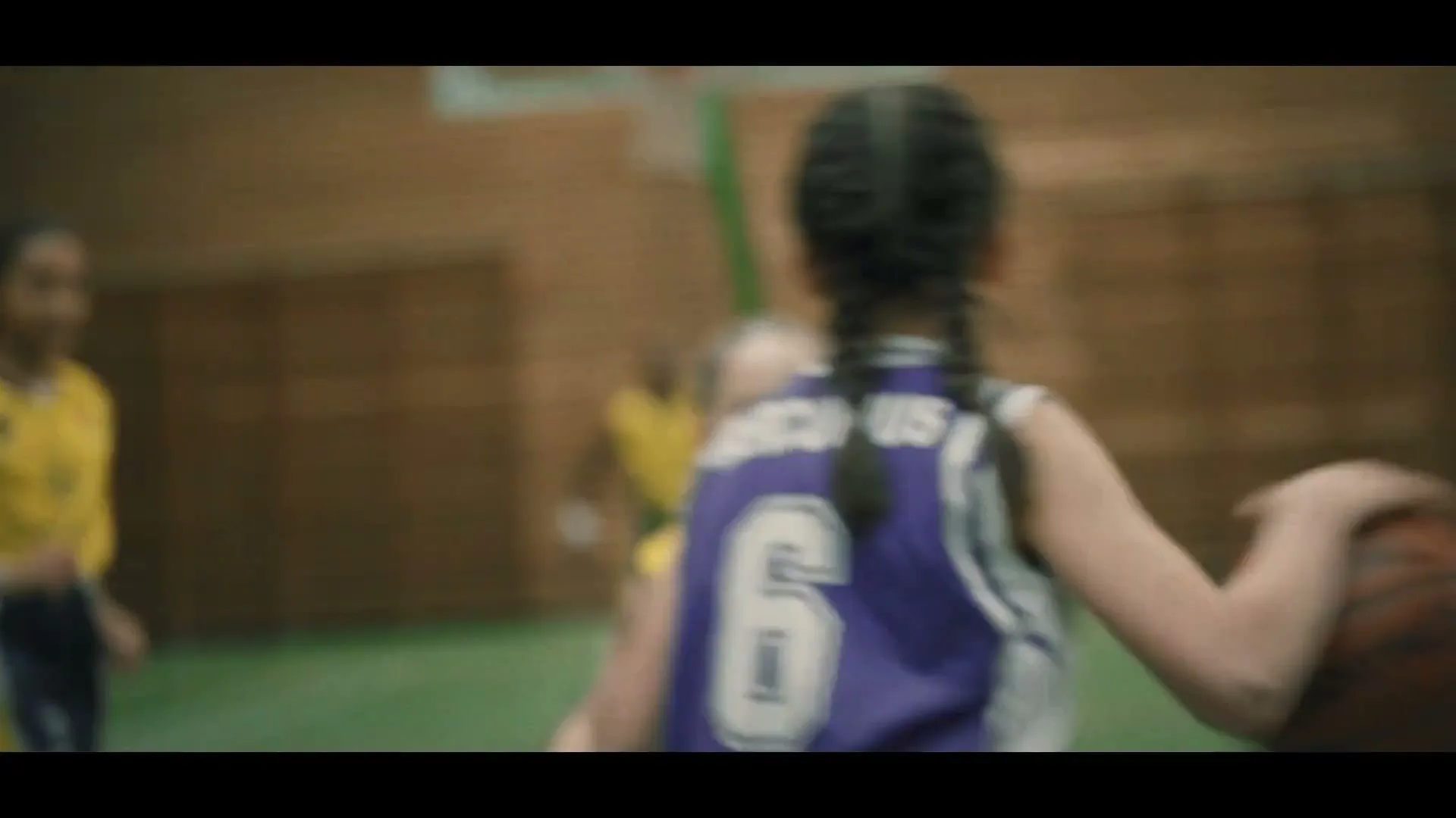 Two young girls play basketball on a court, one in a purple jersey dribbles while another in a yellow jersey defends, with spectators in the background.