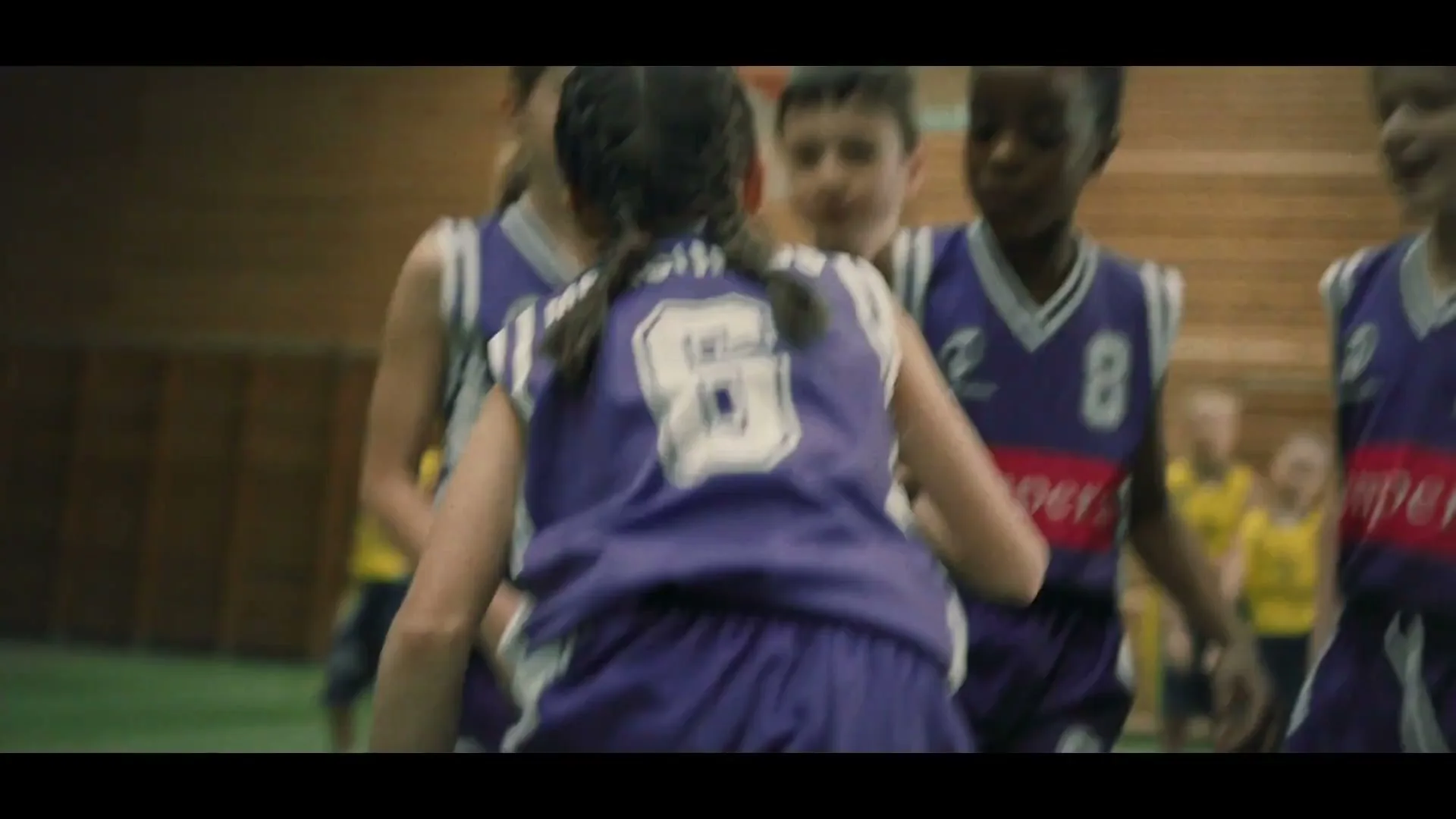 A young girl with braided hair in a purple basketball jersey huddles with her teammates on a basketball court.
