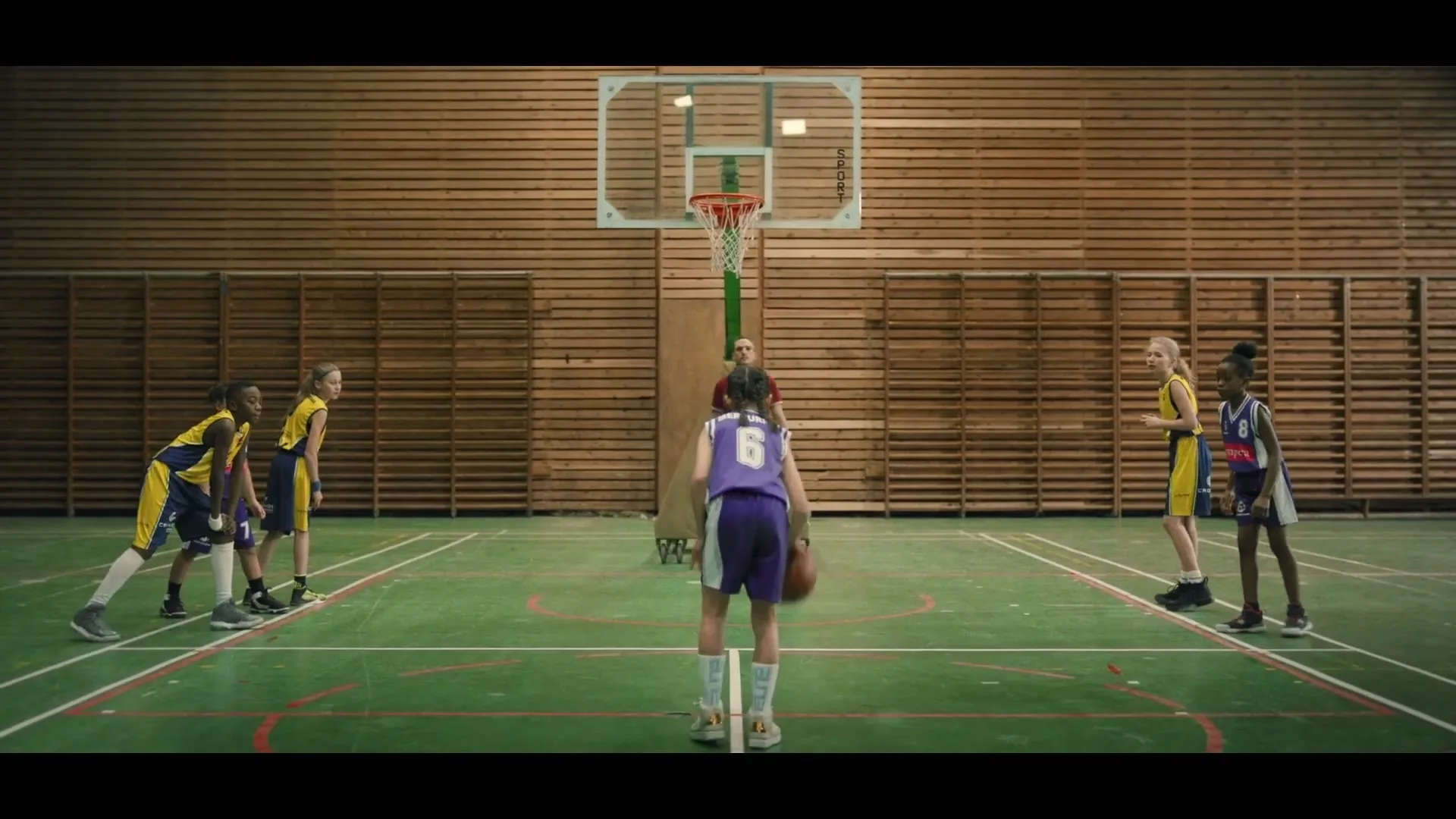 A young girl in a purple basketball uniform dribbles a basketball on a court, facing away from the camera, with other children and an adult coach watching.