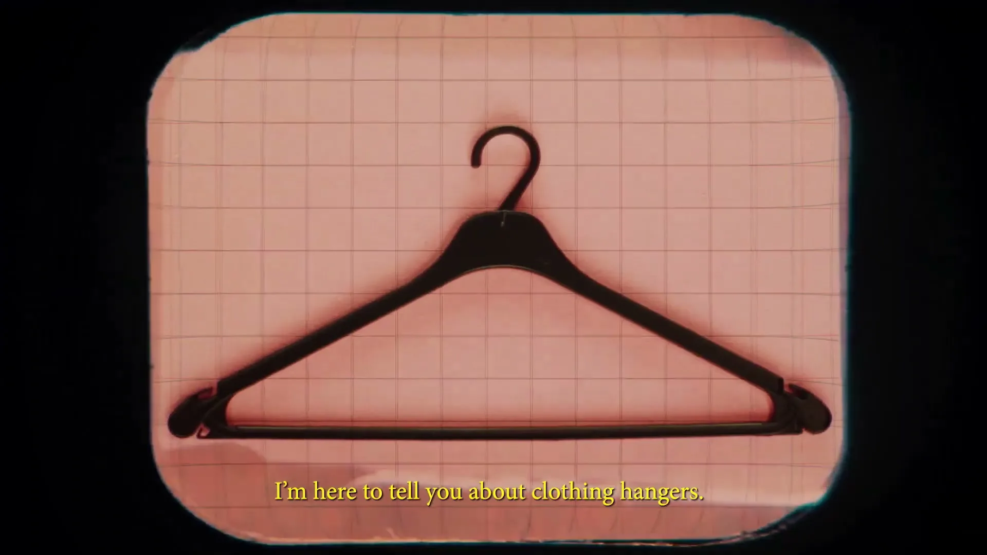 A black plastic clothing hanger is shown against a pink grid background, with text overlayed at the bottom of the screen.