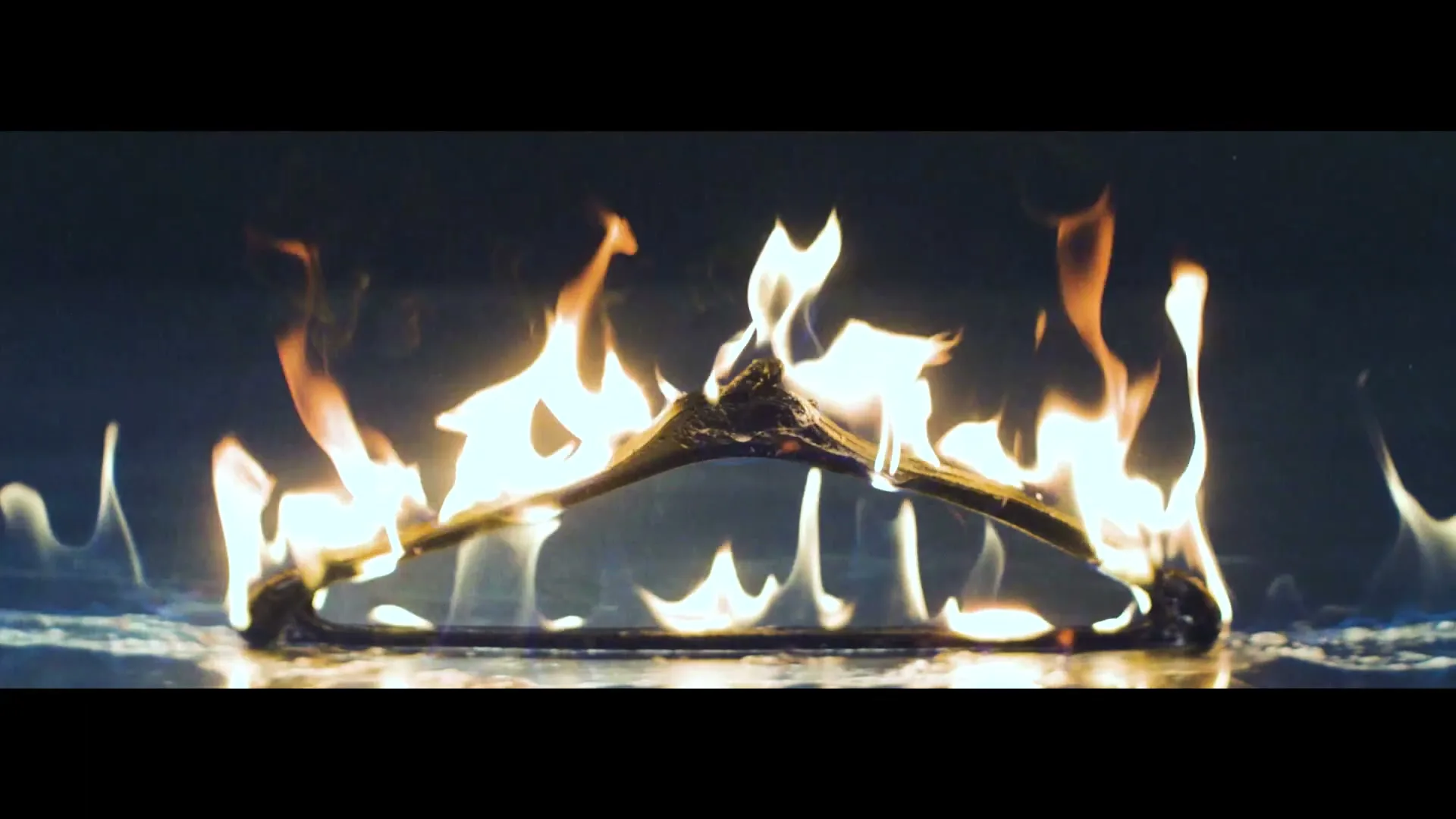 A plastic clothes hanger burns on a dark surface with bright orange and yellow flames against a dark background.