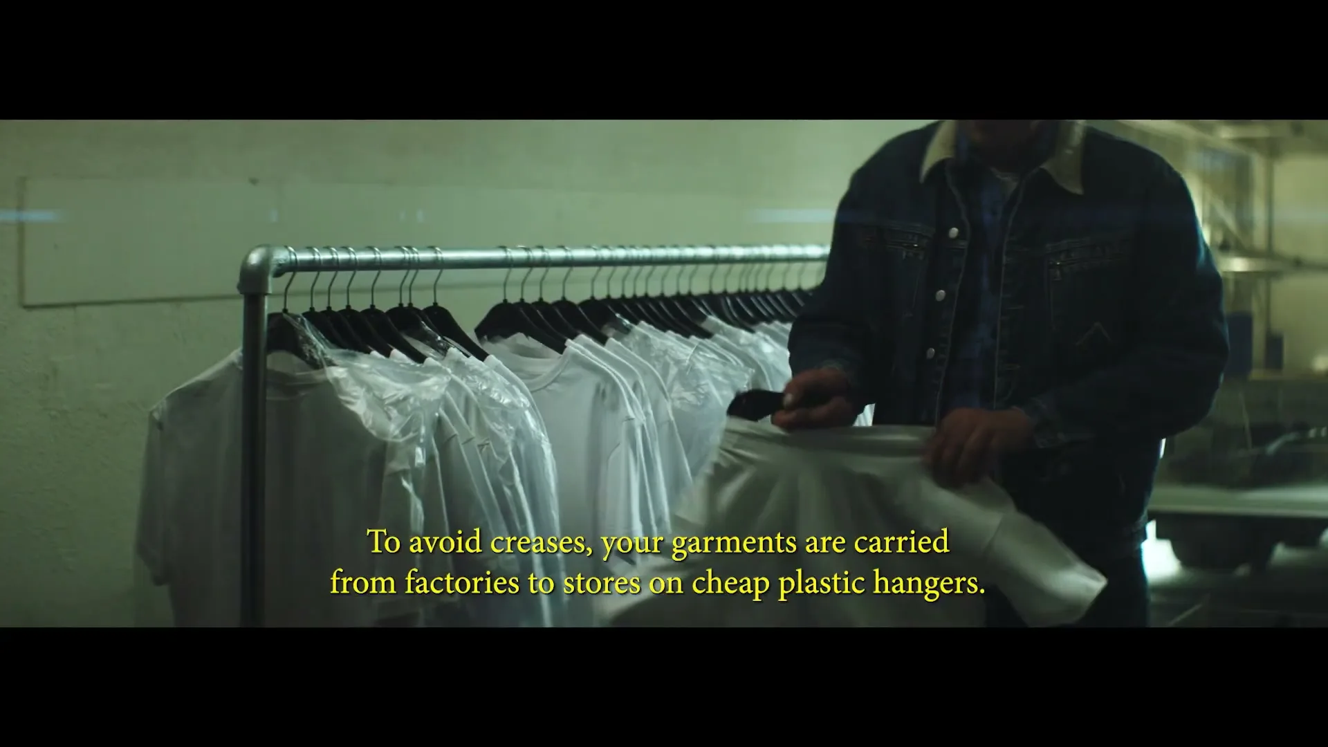 A worker in a denim jacket hangs white t-shirts on a clothing rack in a dimly lit factory or warehouse, with text overlay about plastic hangers.