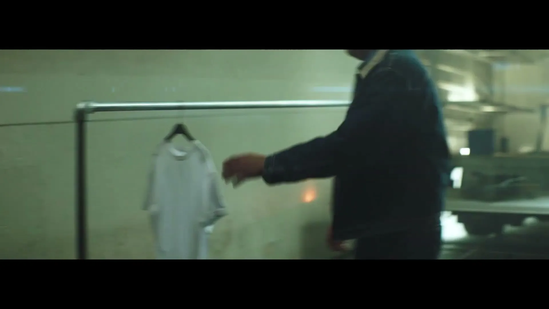 A person in a dark jacket hangs a white t-shirt on a metal rack in a dimly lit industrial setting, then walks away.