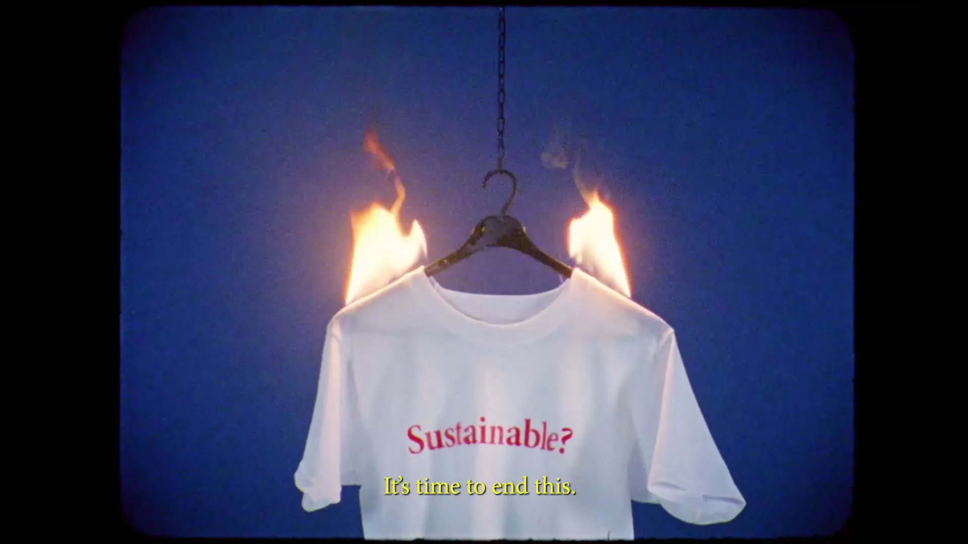 A white t-shirt with the word "Sustainable?" printed on it hangs on a hanger and is on fire against a blue background.