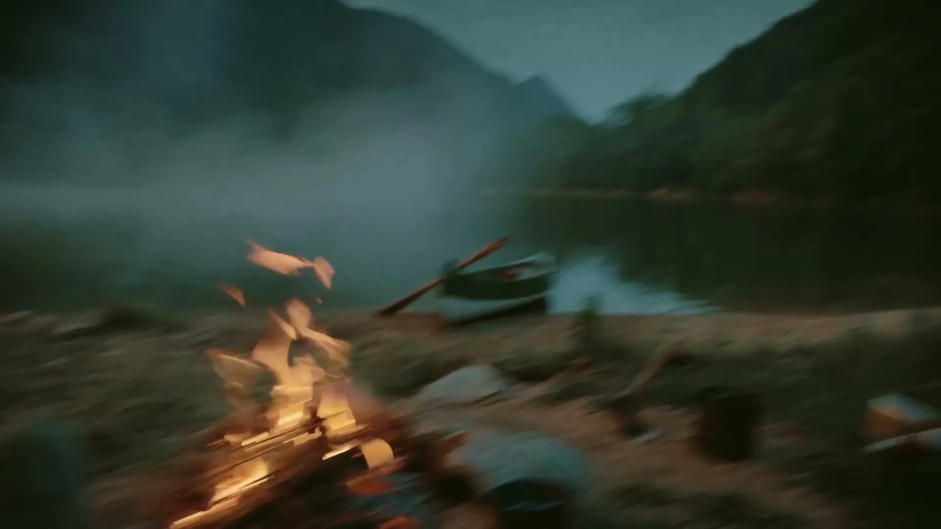 A person's feet are seen resting by a campfire on a lakeshore, with a canoe and mountains in the background, then the camera pans to a tent.