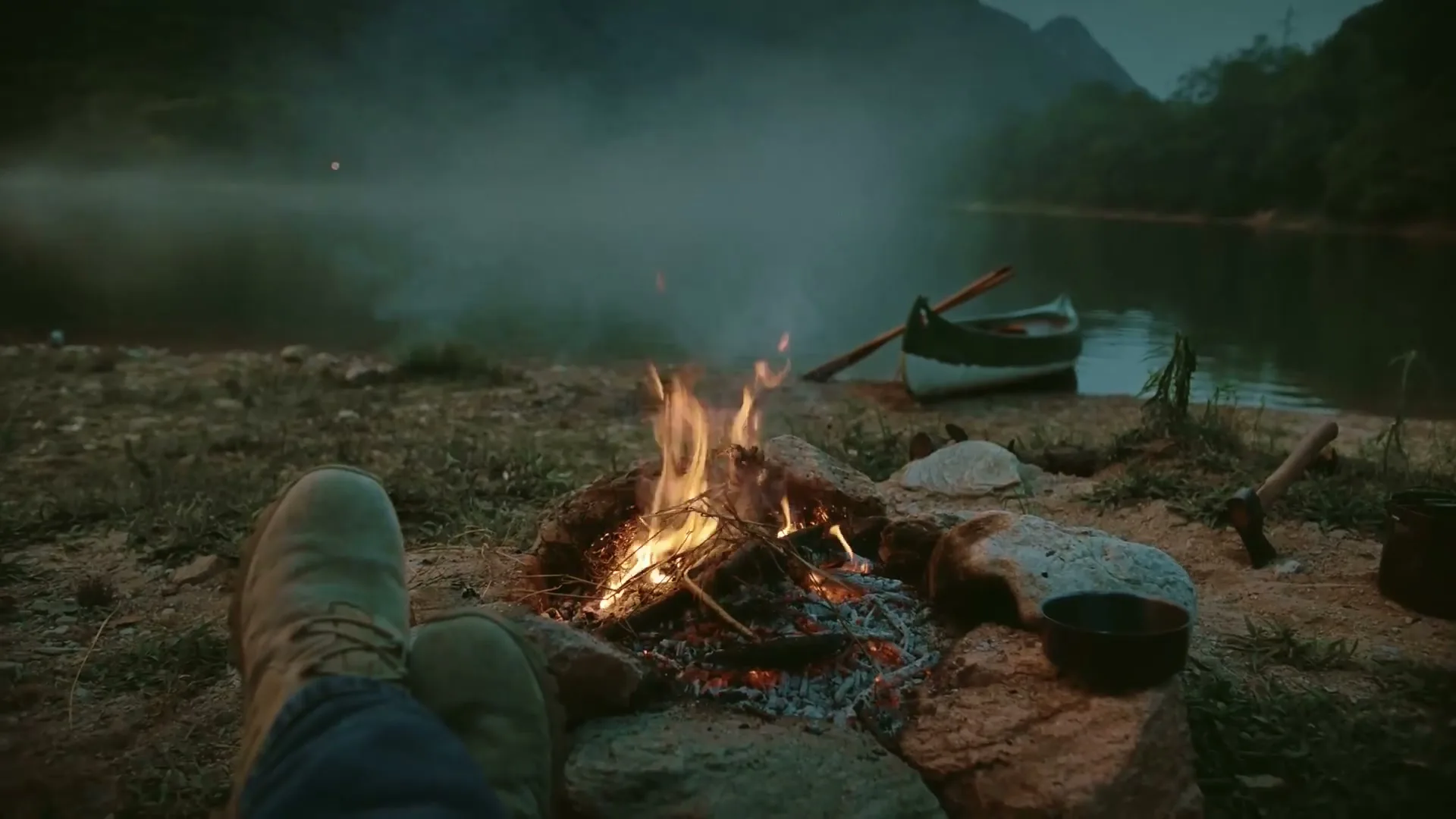 A person's feet are propped up by a campfire, with a canoe on a misty lake and mountains in the background at dusk.