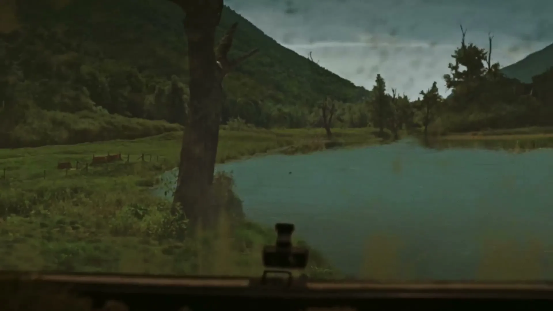 A wide shot from inside a vehicle shows a lake surrounded by green hills and trees, with a dark, blurry foreground, as the vehicle moves quickly.