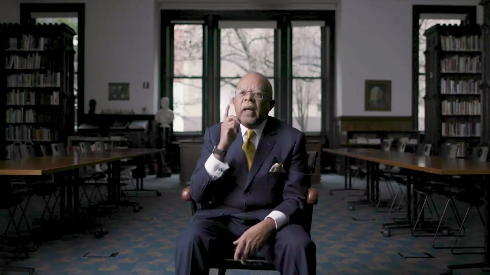 A distinguished older Black man in a suit and tie sits in a chair, gesturing as he speaks in a library with large windows and bookshelves.
