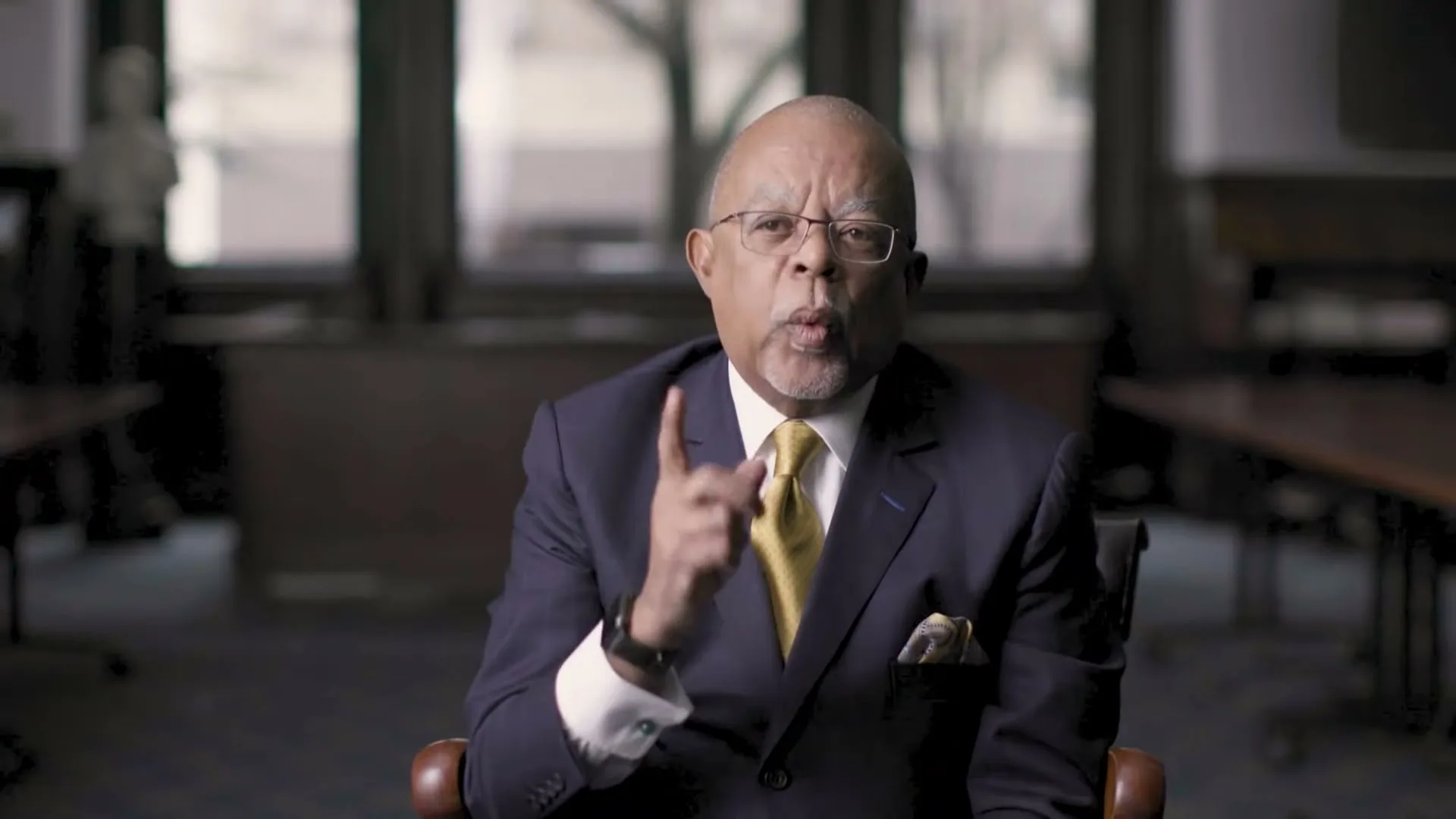 A distinguished older Black man in a suit and glasses speaks directly to the camera, gesturing with his finger in a dimly lit room.