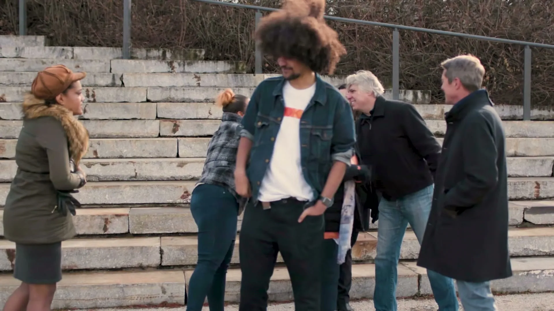 A diverse group of people, including a man with a large afro, gather on concrete steps outdoors, greeting each other.