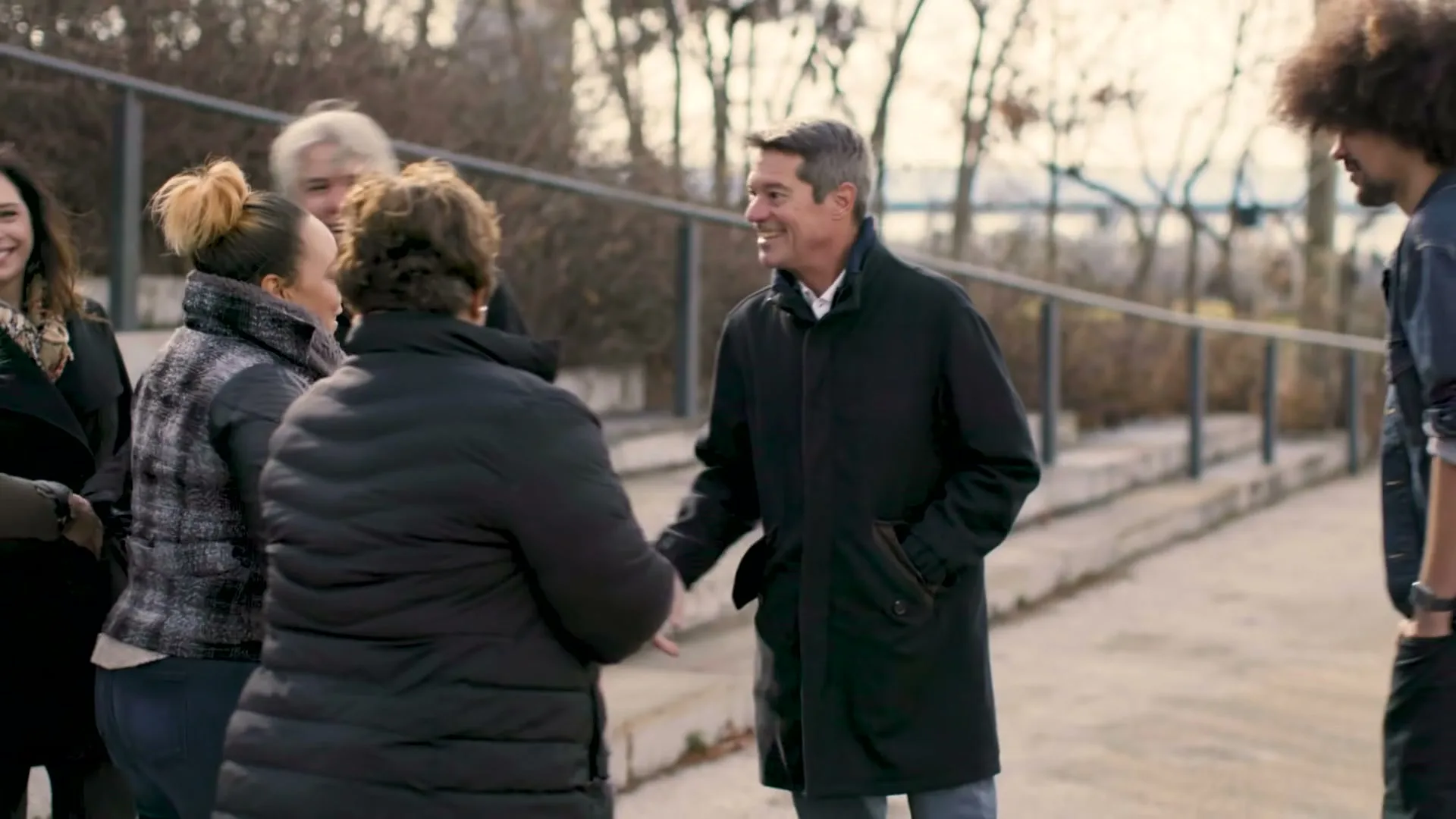 A diverse group of people gather outdoors, with a man in a dark coat shaking hands with a woman in a puffer jacket, all smiling.