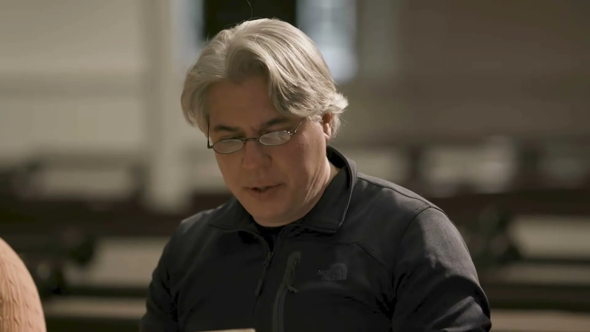 A man with gray hair and glasses, wearing a black jacket, looks down and speaks in a church.