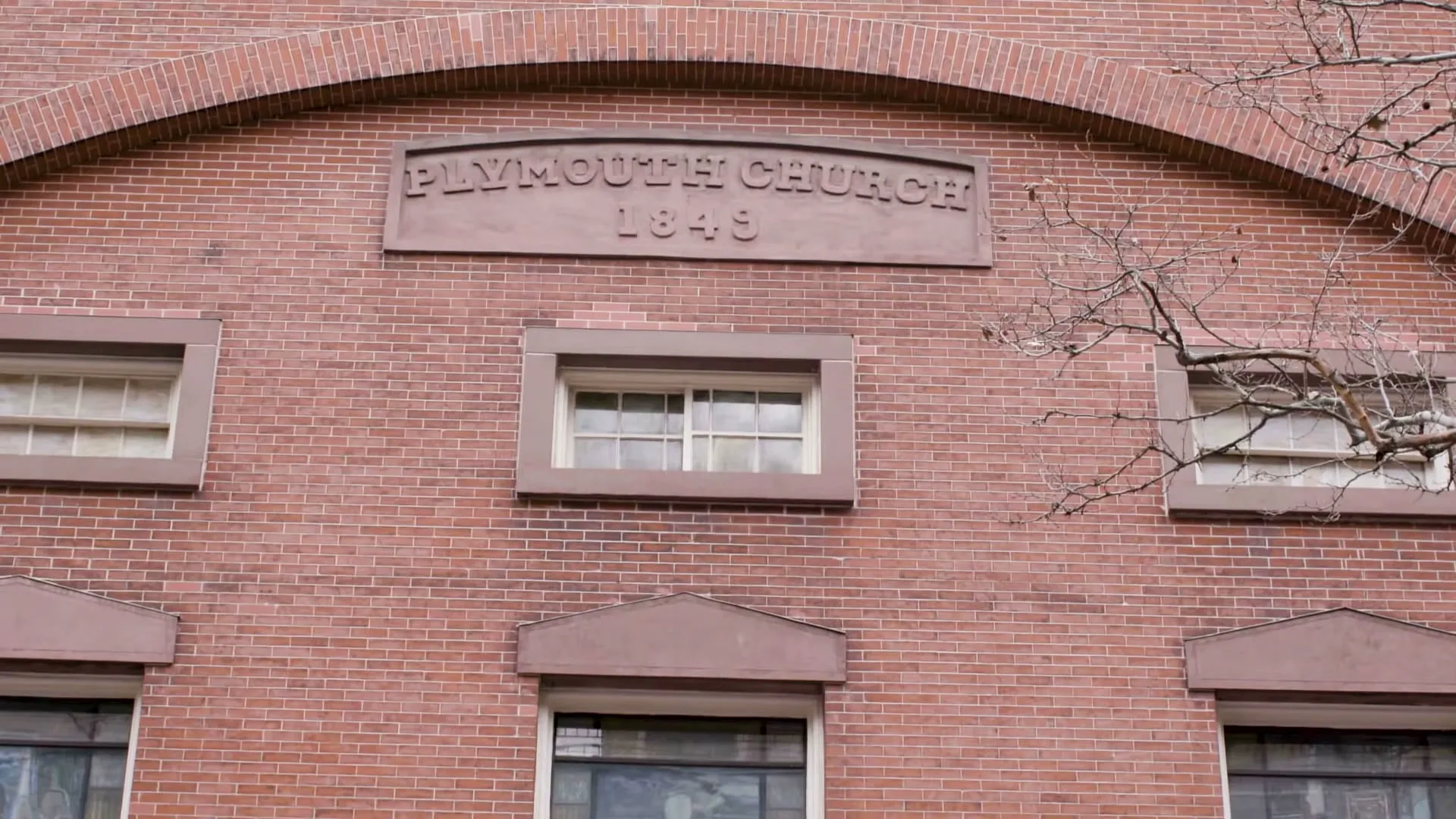 A close-up shot of the brick facade of Plymouth Church, with "PLYMOUTH CHURCH 1849" inscribed above a central window, and bare tree branches on the right.