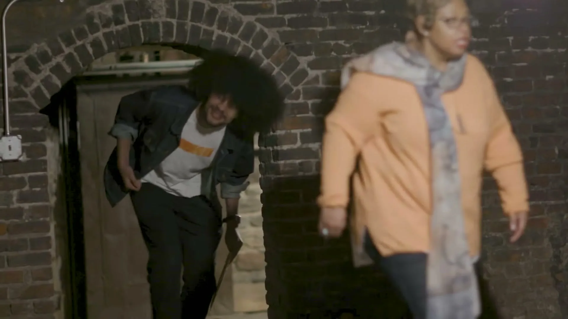 A man with an afro and a woman in an orange cardigan walk through a brick archway in a dimly lit tunnel.