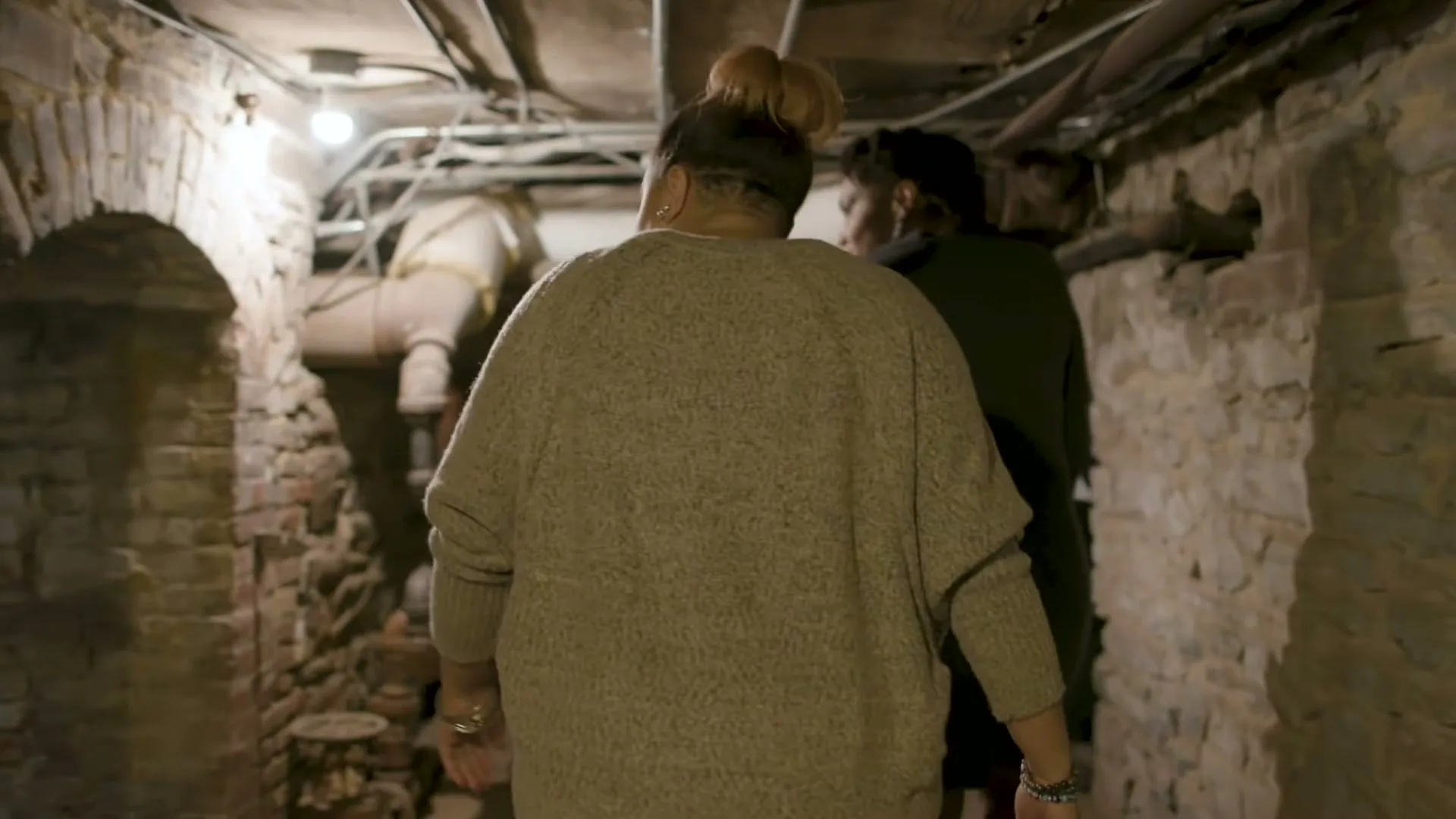 Two women walk through a dimly lit, narrow brick and stone basement with exposed pipes overhead, exploring the old structure.