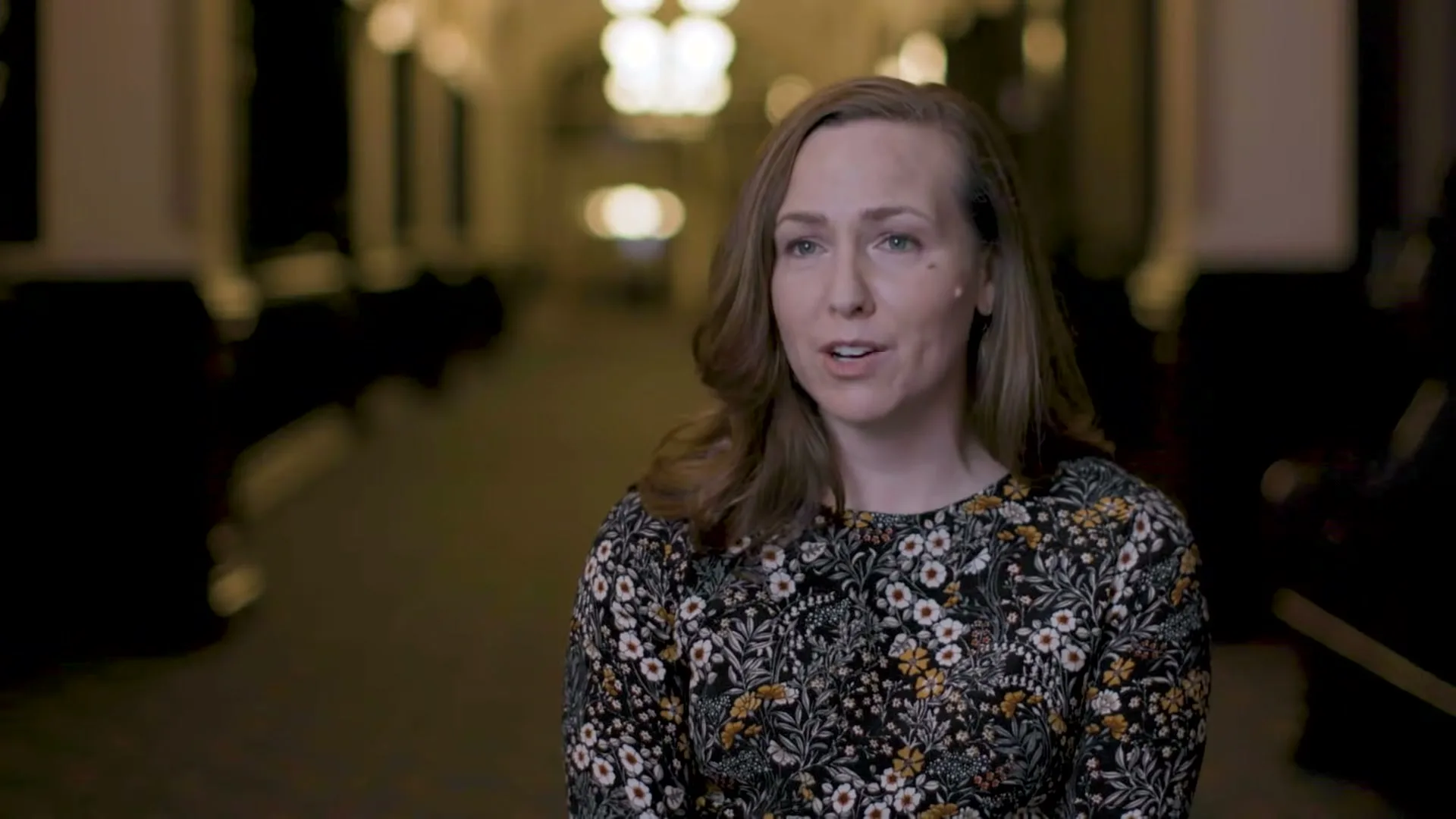 A woman with brown hair and a floral top speaks directly to the camera in a dimly lit, ornate interior hallway.