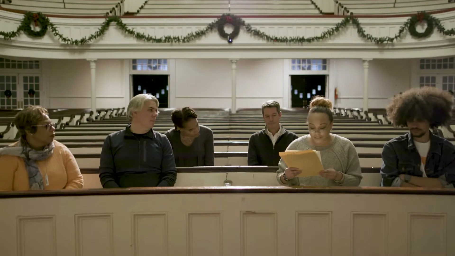 A diverse group of people sit in church pews, listening intently as a woman reads from a document in a church decorated for Christmas.