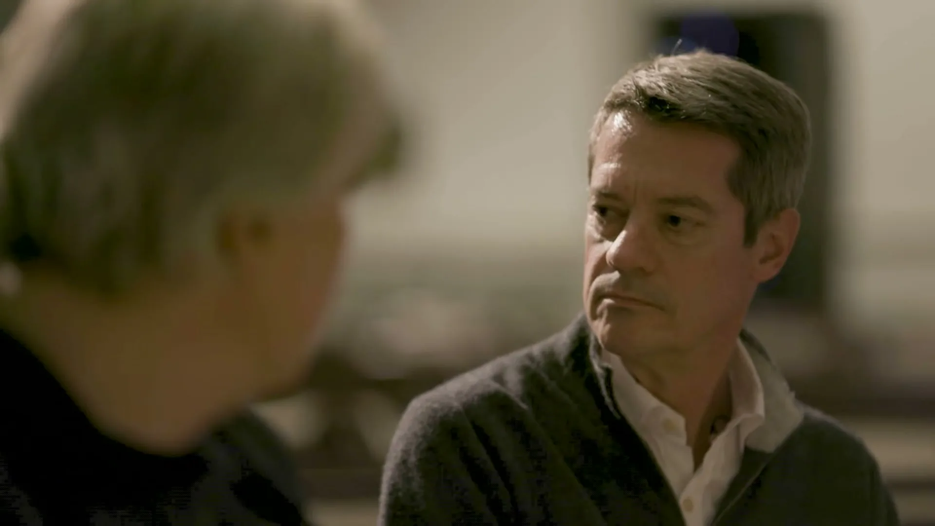 A man with grey hair and a white shirt looks intently at another person, whose back is to the camera, in an indoor setting.