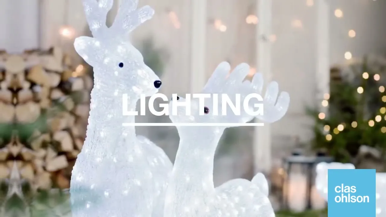 Two white, glowing reindeer decorations stand in a festive outdoor setting with firewood and string lights in the background.