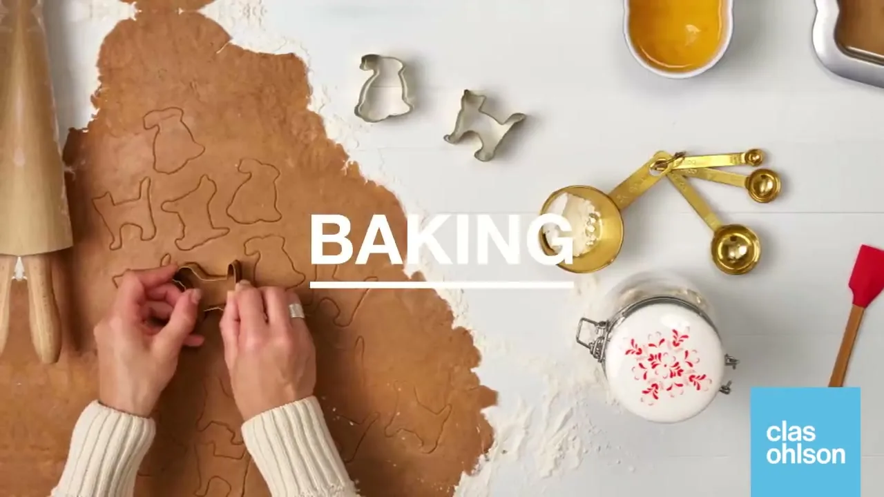 A person's hands cut out gingerbread cookies from dough on a white table with baking ingredients, with the word "BAKING" on screen.