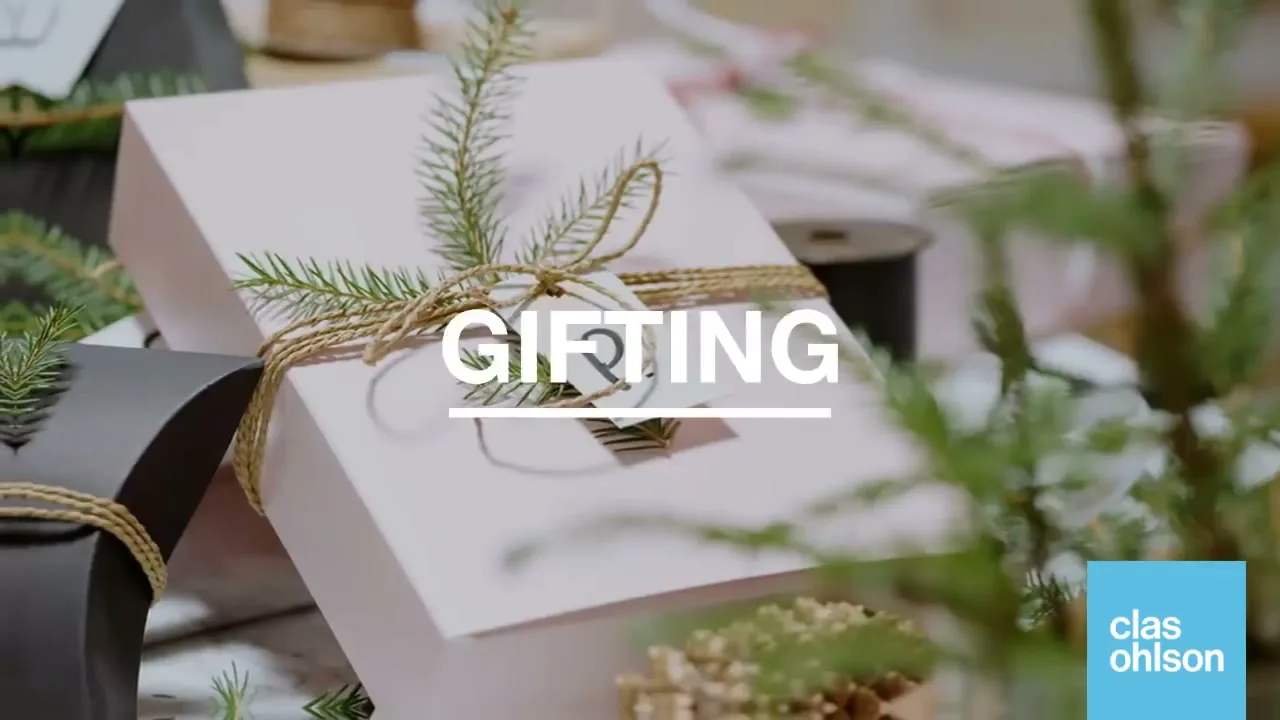 A close-up shot of a beautifully wrapped pink gift box with a sprig of pine and a tag, surrounded by other gifts and greenery, with the word "GIFTING" overlaid.